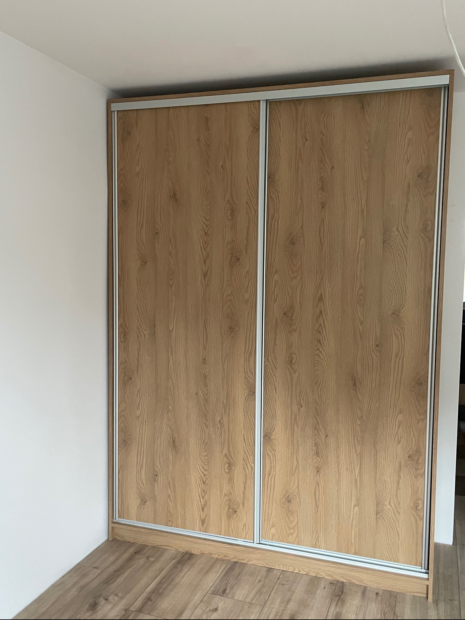 Szafa wnękowa z przesuwnymi drzwiami o wzorze jasnego drewna, z metalowymi prowadnicami, dopasowana do rogu pokoju z drewnianą podłogą.