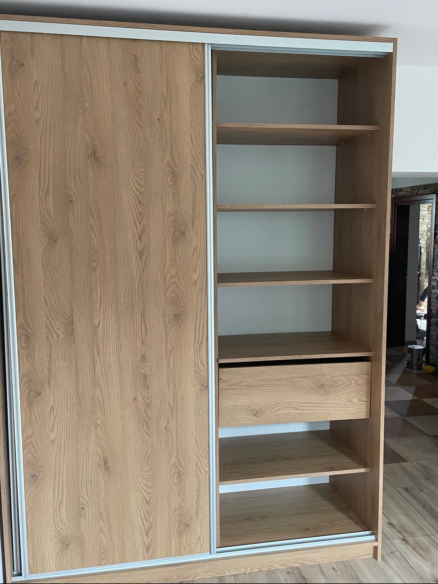 Jasna, drewnopodobna szafa przesuwna z otwartą częścią regałową, widoczna szuflada i puste półki, prezentacja konstrukcji i wykończenia.