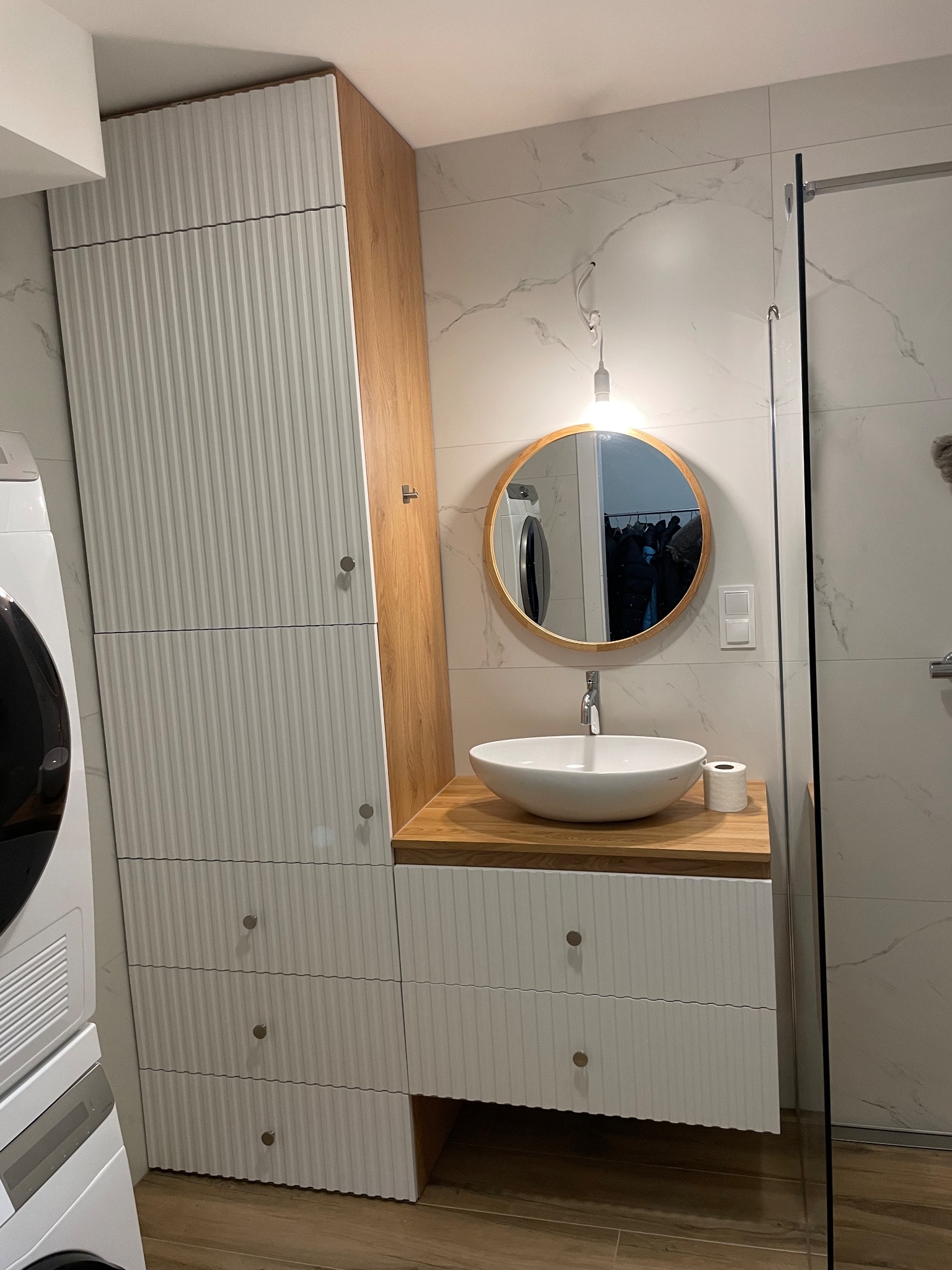 Nowoczesna szafa łazienkowa na wymiar z białymi, ryflowanymi frontami i drewnianym blatem, obok pralka, lustro w okrągłej ramie i kabina prysznicowa.