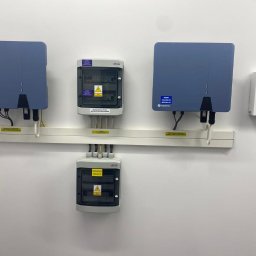 Electric Sp. jawna - Dwa niebieskie inwertery fotowoltaiczne Solplanet i dwie szare skrzynki rozdzielcze ETI zamontowane na białej ścianie na białej szynie montażowej, z widocznymi kablami i naklejkami ostrzegawczymi.