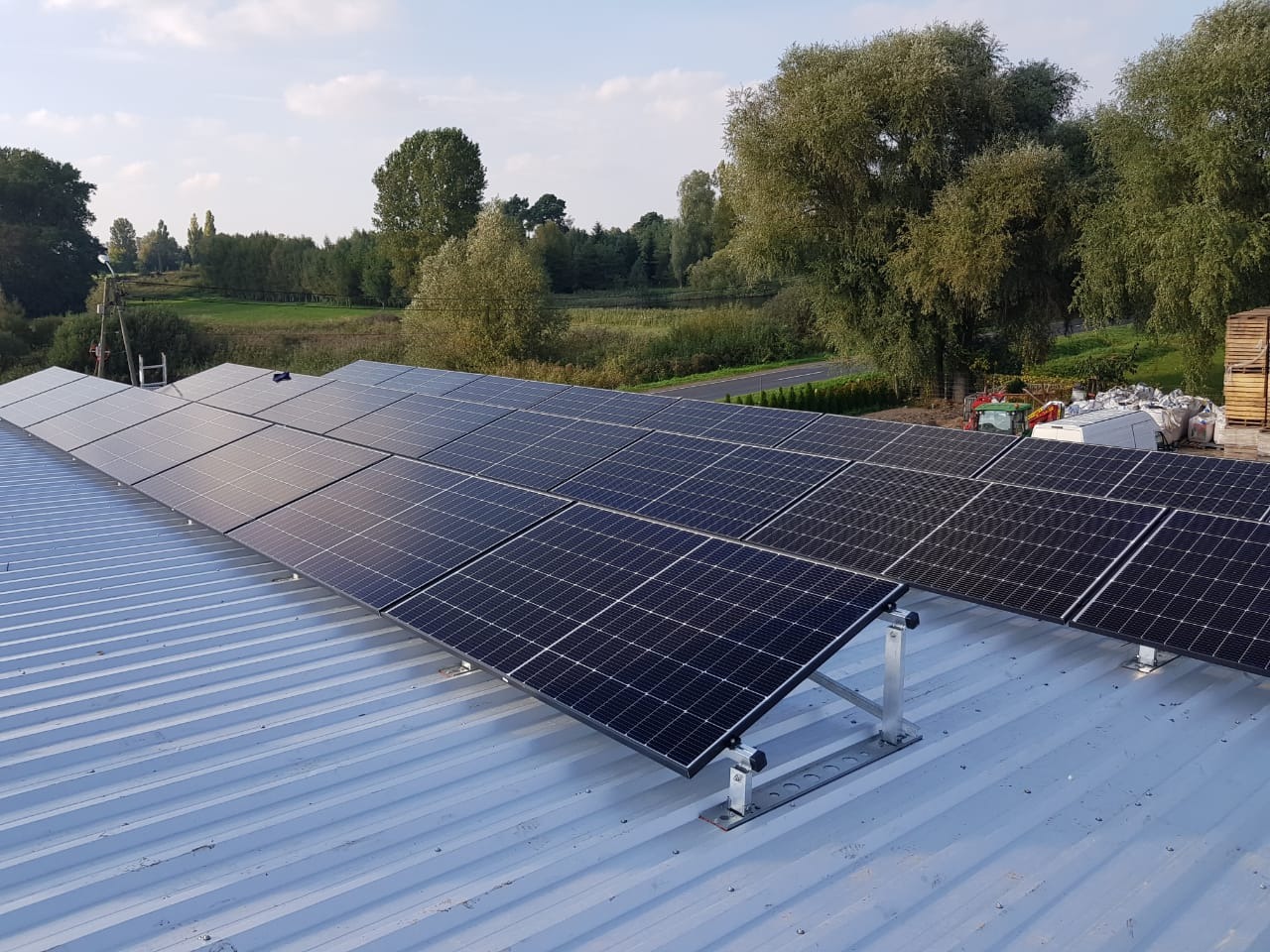 Panele słoneczne zamontowane na blaszanym dachu z widokiem na zielone drzewa i pole w tle. Widoczne metalowe wsporniki mocujące panele.