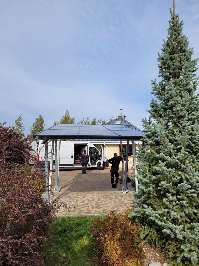 Montaż konstrukcji z panelami słonecznymi na zewnątrz budynku, widoczny biały samochód dostawczy i dwóch instalatorów w trakcie pracy, z boku duży świerk.