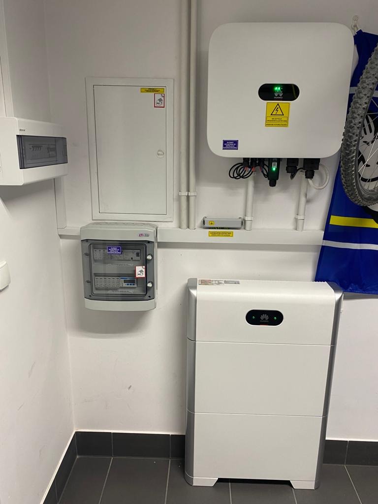 Biała bateria magazynu energii Huawei obok szafki z bezpiecznikami i inwertera fotowoltaicznego na białej ścianie z szarą podłogą, widoczny fragment roweru.