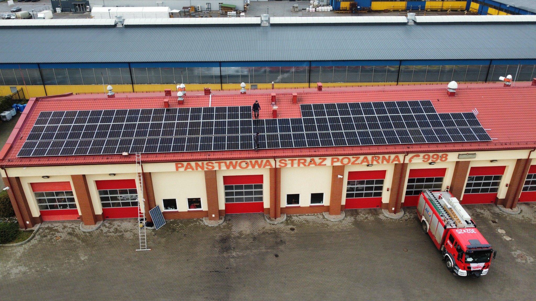 Widok z góry na remizę strażacką z panelami słonecznymi na czerwonym dachu, dwaj mężczyźni na dachu, czerwony wóz strażacki zaparkowany przed budynkiem.
