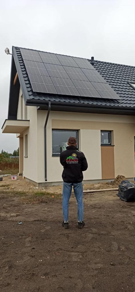 Dom jednorodzinny z zamontowanymi panelami słonecznymi na dachu, obserwowany przez mężczyznę w kurtce z logo firmy elektrycznej. Widoczne świeżo pomalowane ściany budynku i niezagospodarowany teren...