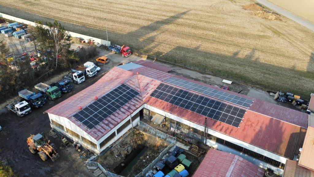 Widok z góry na budynek z czerwonym dachem, na którym zamontowano panele słoneczne. Obok budynku wykopany dół, a w tle widać zaparkowane ciężarówki i pole uprawne.