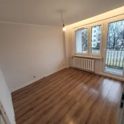 Pusty pokój po remoncie z brązowymi panelami podłogowymi, białymi ścianami, grzejnikiem pod oknem i gołą żarówką zwisającą z sufitu.