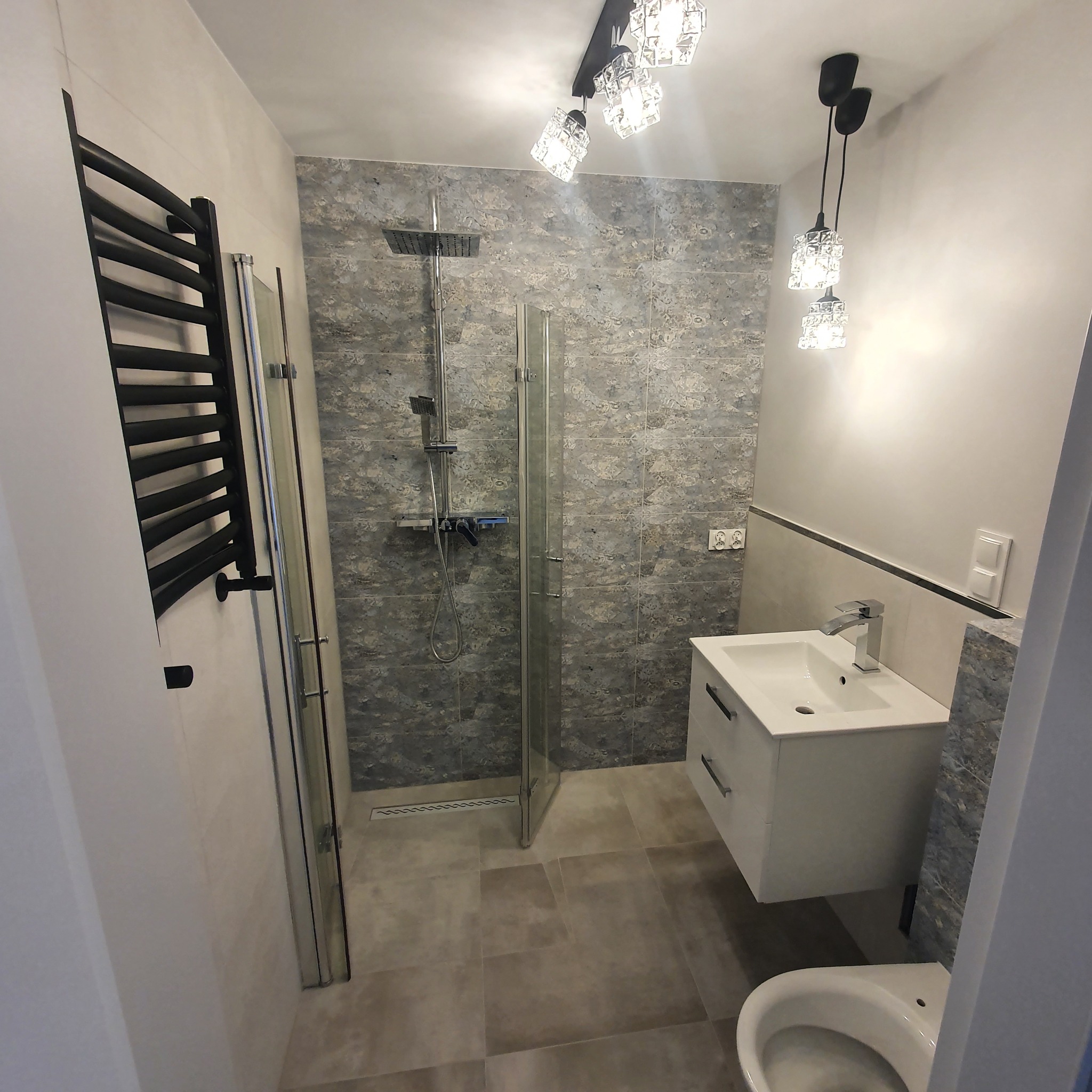 Nowoczesna łazienka z szarymi płytkami imitującymi kamień, kabiną prysznicową typu walk-in, białą szafką z umywalką i grzejnikiem drabinkowym w kolorze czarnym. Oświetlenie stanowią kryształowe...