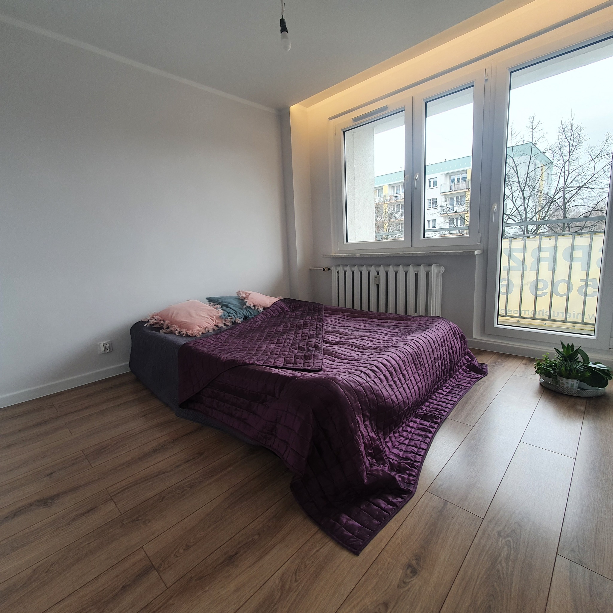 Pokój z drewnianą podłogą, szarym łóżkiem z fioletową narzutą i dekoracyjnymi poduszkami, widok na budynek przez okno z grzejnikiem i roślinami w metalowej tacy.