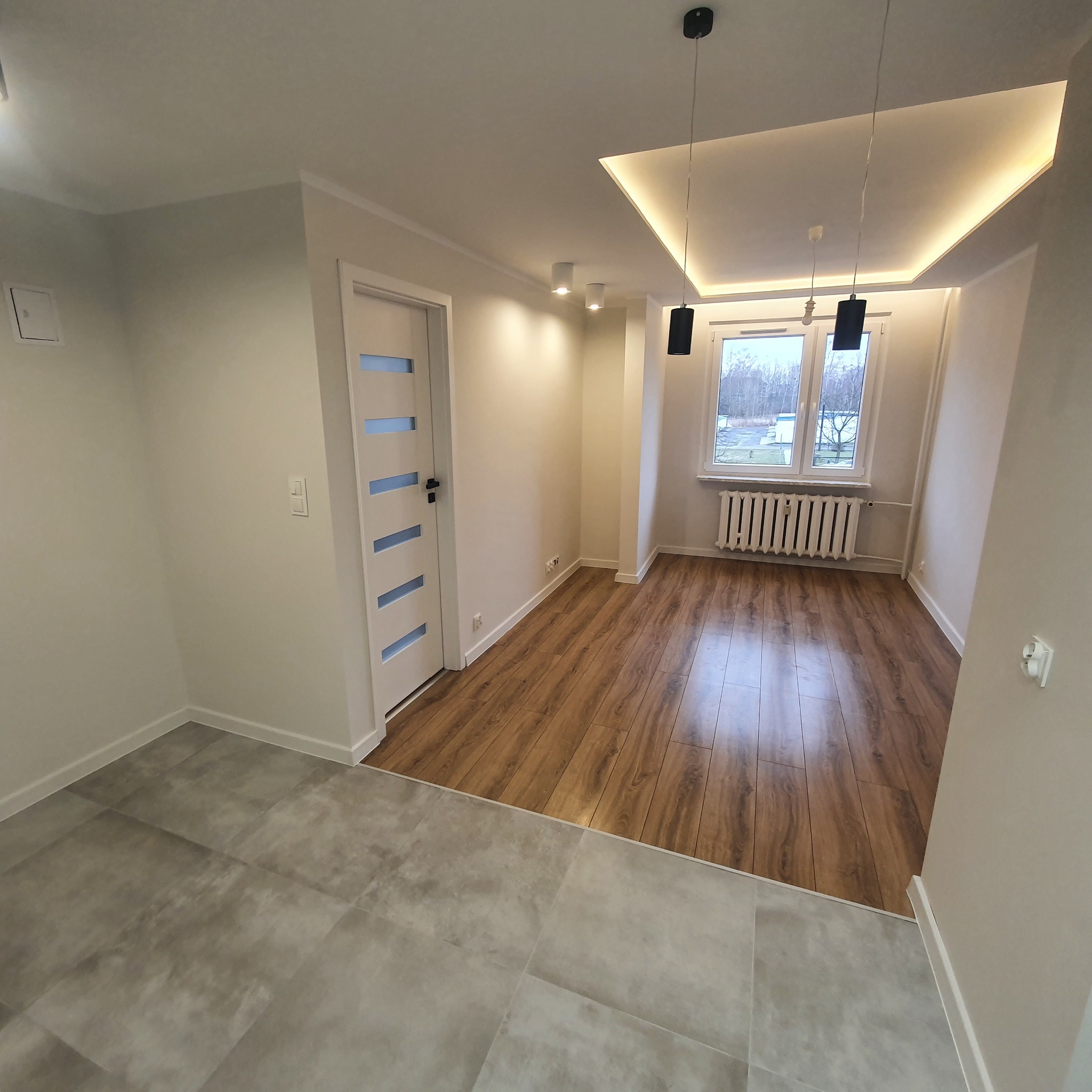 Wykończone wnętrze: szara podłoga z płytek przechodzi w drewnopodobne panele, białe ściany, drzwi z przeszkleniami, grzejnik pod oknem i nowoczesne oświetlenie sufitowe.