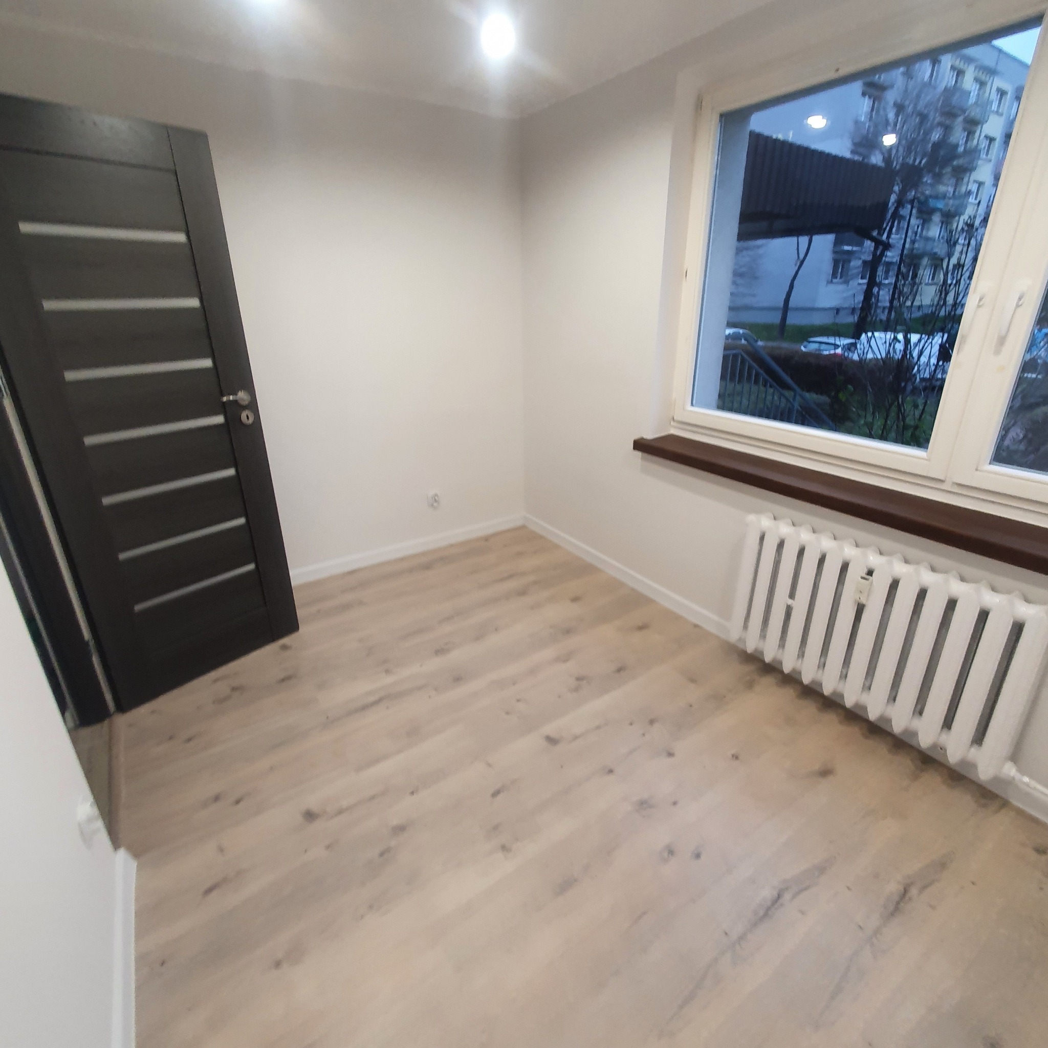 Jasna podłoga z paneli ułożona w pokoju z białymi ścianami, ciemnymi drzwiami z poziomymi wstawkami i białym kaloryferem pod oknem z widokiem na blok.