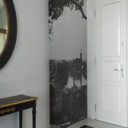 Szafa wnękowa z nadrukiem czarno-białego zdjęcia panoramy miasta z mostem i drzewem, umieszczona w przedpokoju z czarno-białą szachownicą na podłodze, obok białych drzwi i konsoli z czarnym blatem...