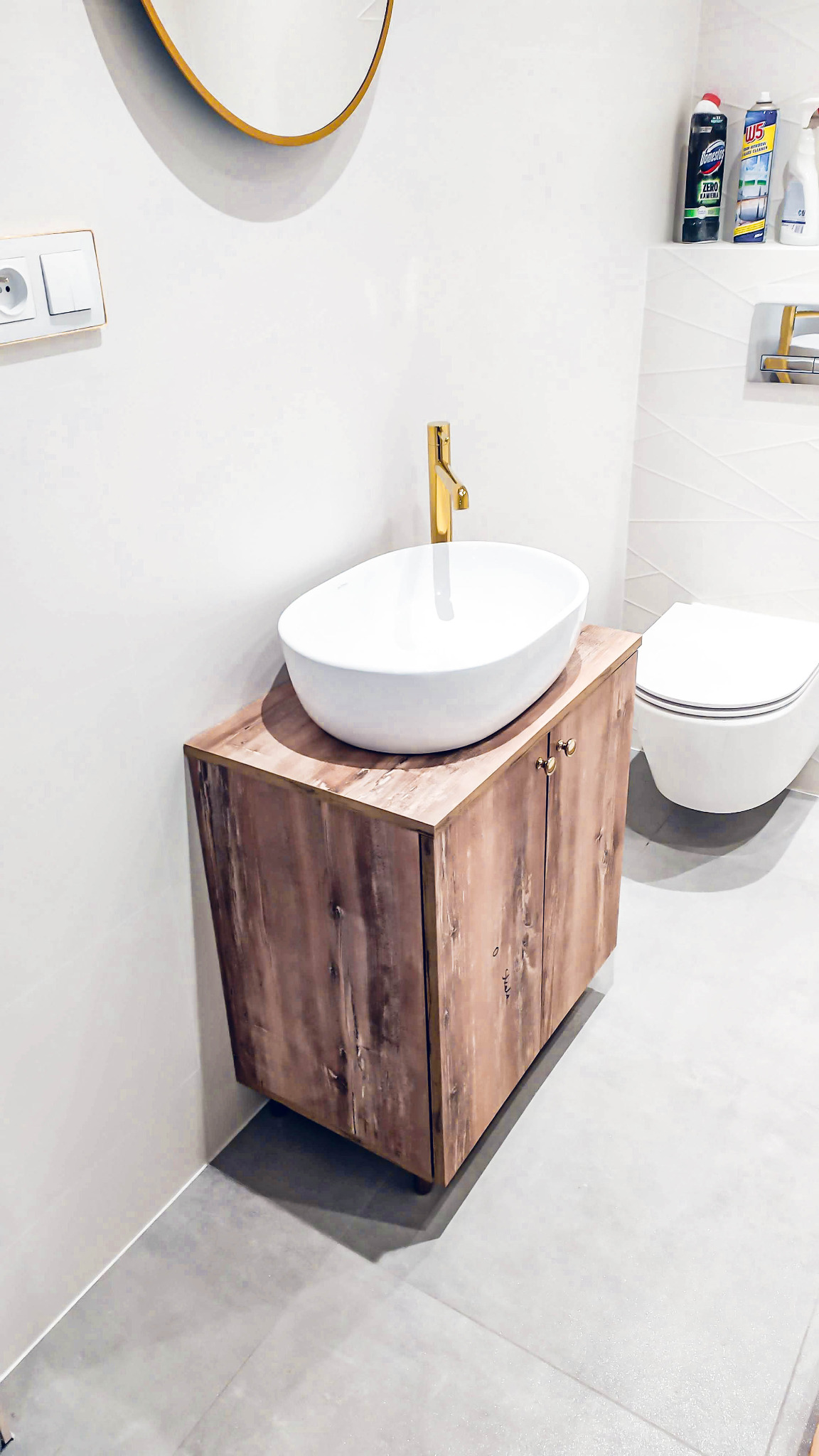 Nowoczesna łazienka z umywalką nablatową w kształcie owalu, złota bateria, szafka pod umywalkę z drewnopodobnym fornirem, okrągłe lustro ze złotą ramą i wisząca toaleta w tle.