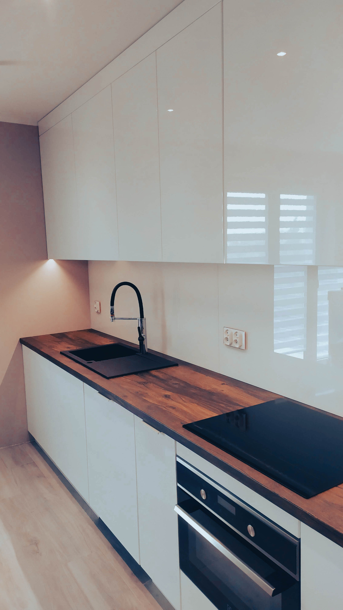 Nowoczesna kuchnia z białymi, błyszczącymi frontami szafek, drewnianym blatem, czarnym zlewem i płytą indukcyjną. Widoczne gniazdka elektryczne na ścianie. Oświetlenie punktowe pod szafkami.