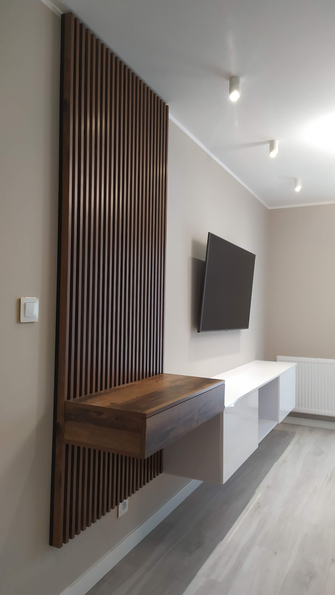 Nowoczesny salon z drewnianą ścianką z pionowych listew, podwieszaną szafką RTV z szufladą i białymi szafkami pod telewizorem.