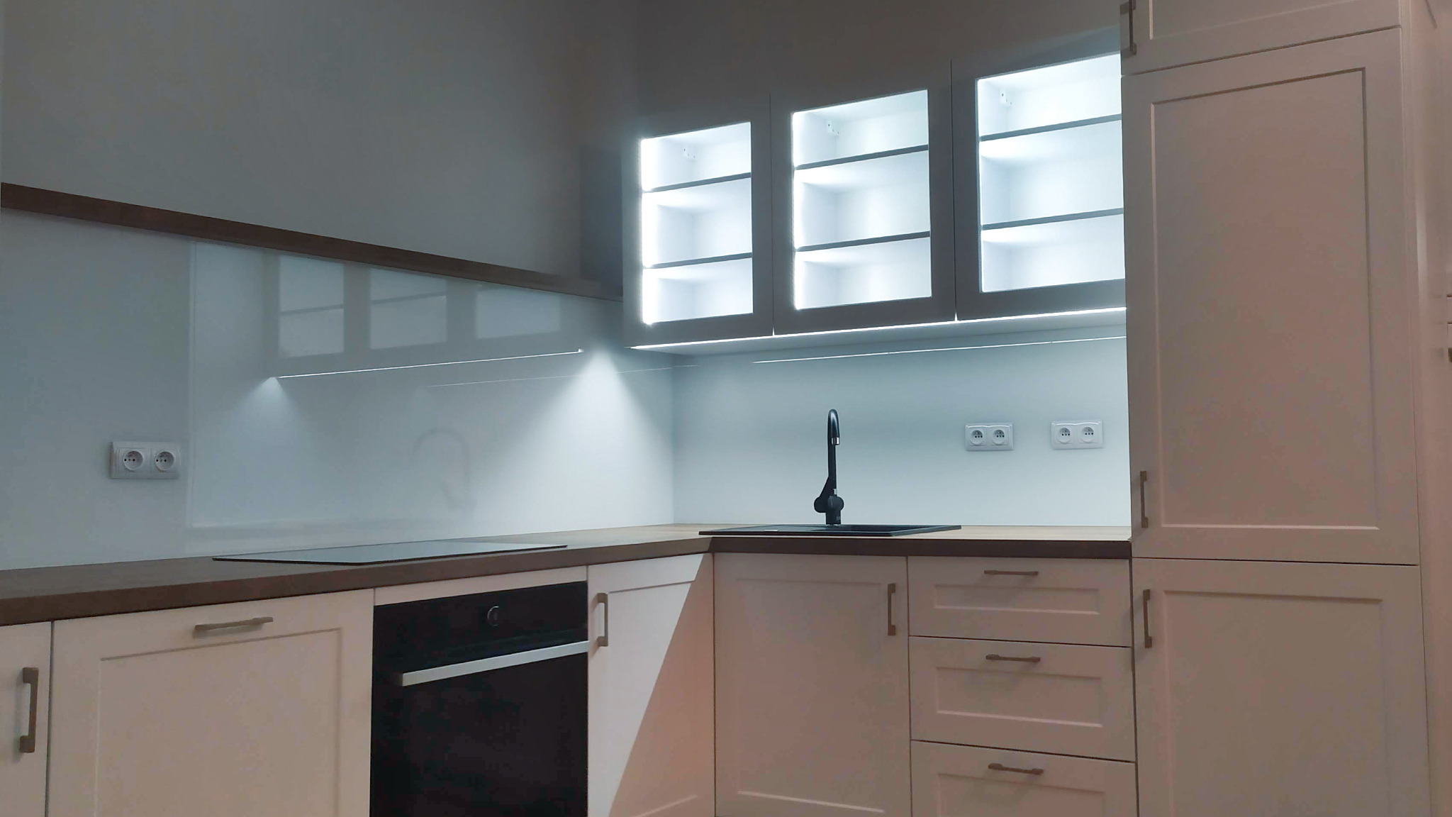 Nowoczesna, jasna kuchnia z białymi szafkami, drewnianym blatem, szklanym panelem nad blatem i oświetleniem LED pod szafkami wiszącymi.