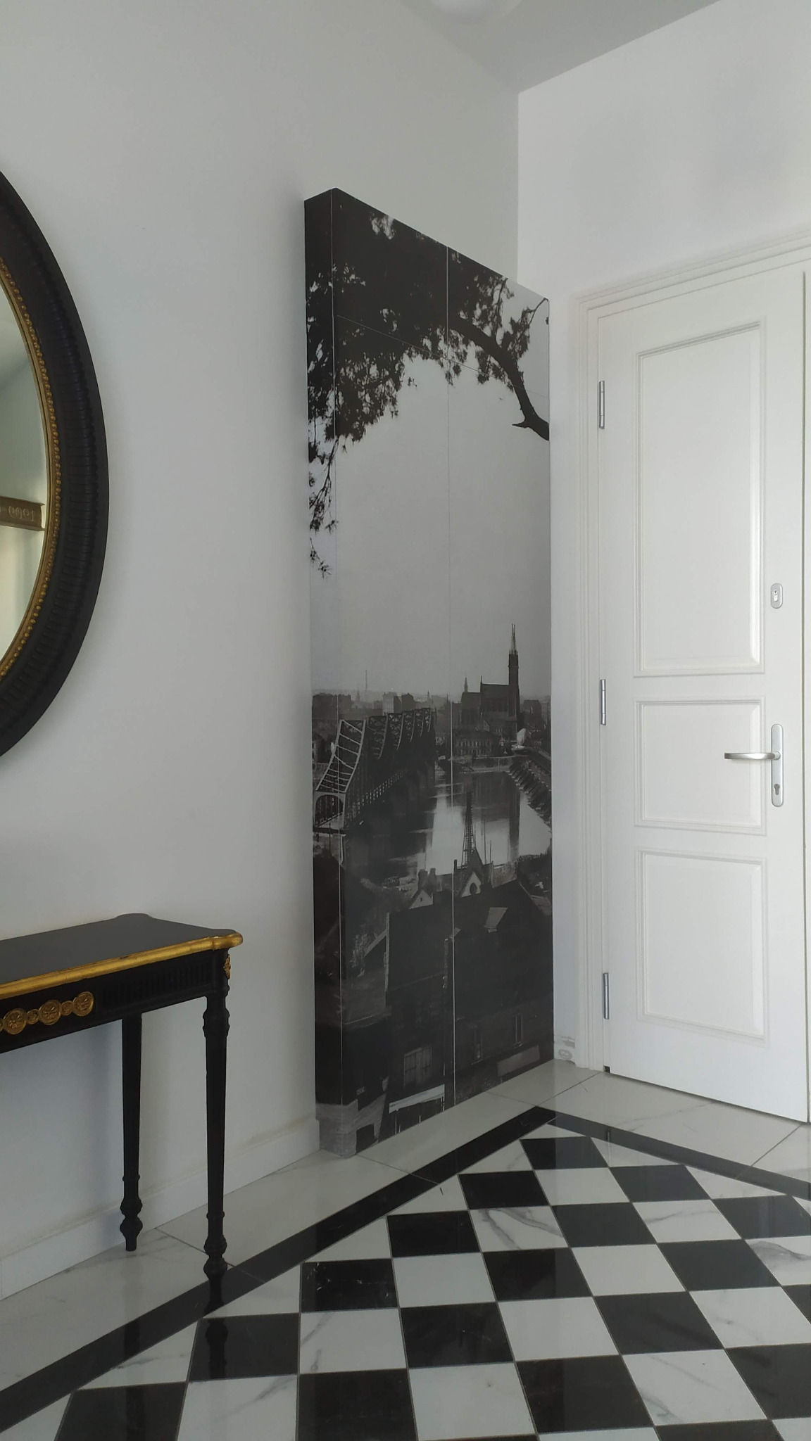 Szafa wnękowa z nadrukiem czarno-białego zdjęcia panoramy miasta z mostem i drzewem, umieszczona w przedpokoju z czarno-białą szachownicą na podłodze, obok białych drzwi i konsoli z czarnym blatem...