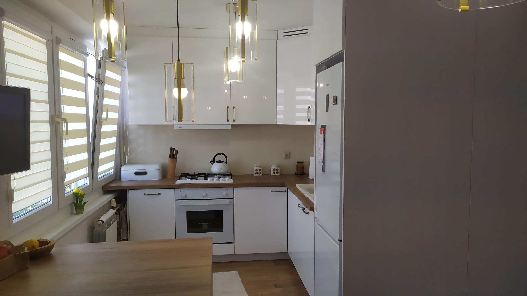 Jasna kuchnia z białymi szafkami, drewnianym blatem i złotymi lampami wiszącymi, widok na okno z żaluzjami poziomymi i drewniany stół