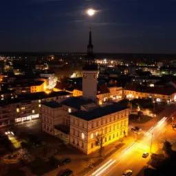 Nocna panorama miasta z ratuszem oświetlonym na pierwszym planie, z smugami świateł samochodowych i księżycem w tle.