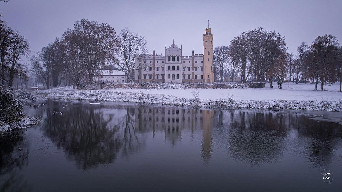 Zimowy krajobraz z zamkiem odbijającym się w wodzie, pokryty śniegiem.