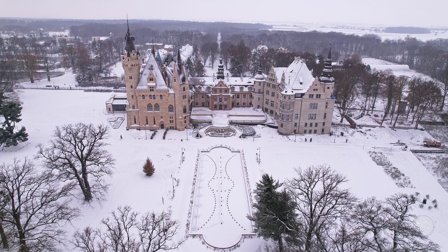 Zimowy widok z lotu ptaka na zabytkowy pałac z wieżami i dziedzińcem pokrytym śniegiem, otoczony ośnieżonymi drzewami i parkiem.