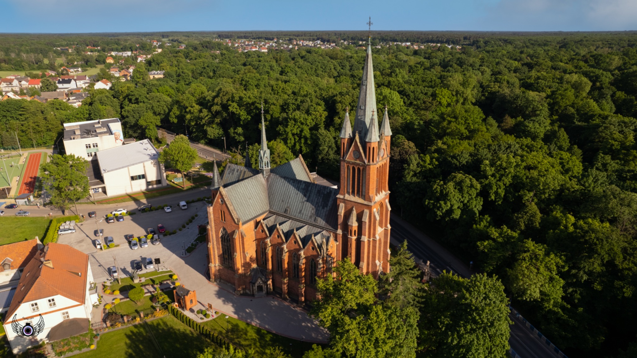 Panorama z lotu ptaka na ceglany kościół z wysoką wieżą, otoczony zielenią drzew i zabudową miejską w słoneczny dzień.