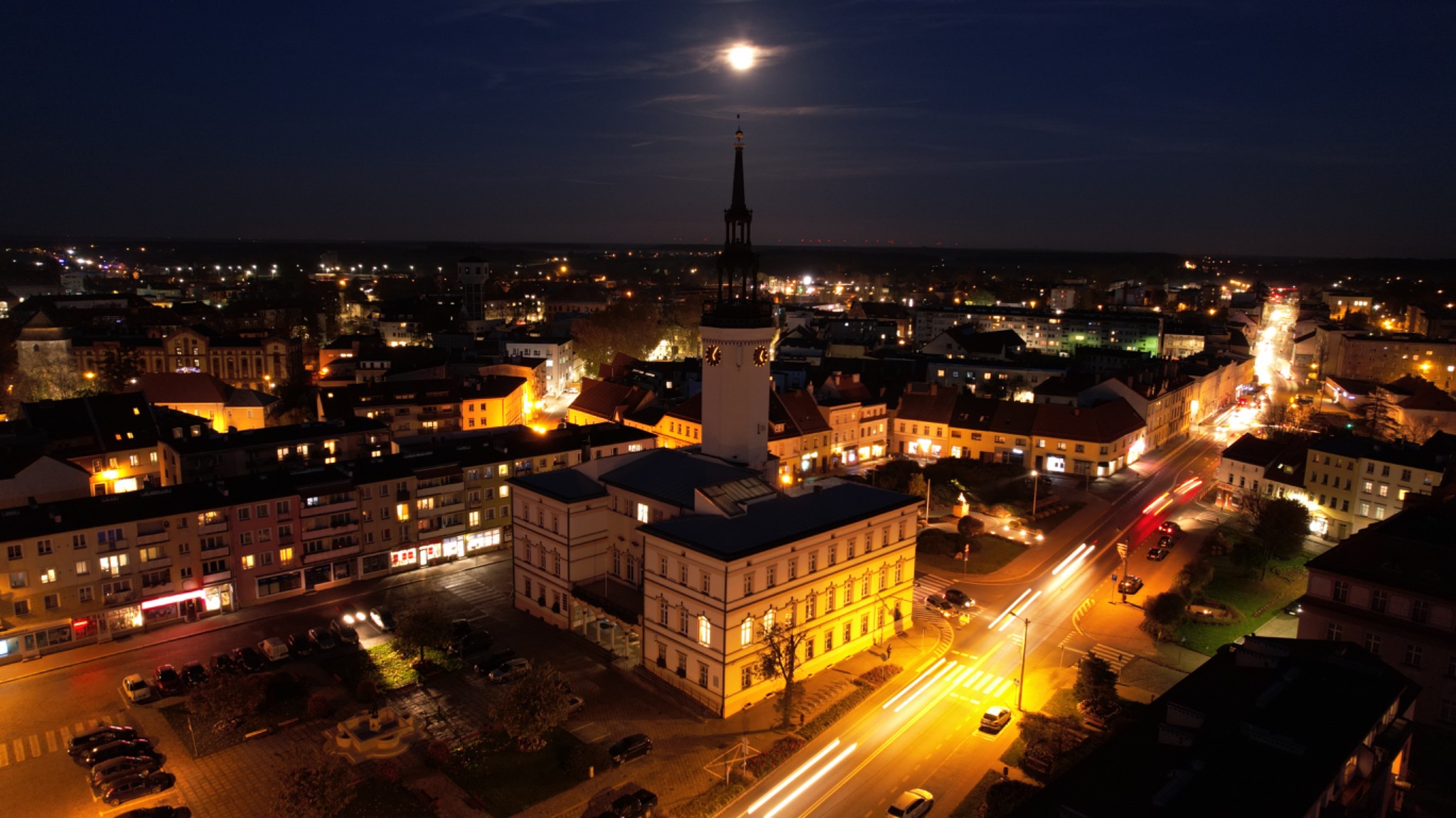 Nocna panorama miasta z ratuszem oświetlonym na pierwszym planie, z smugami świateł samochodowych i księżycem w tle.