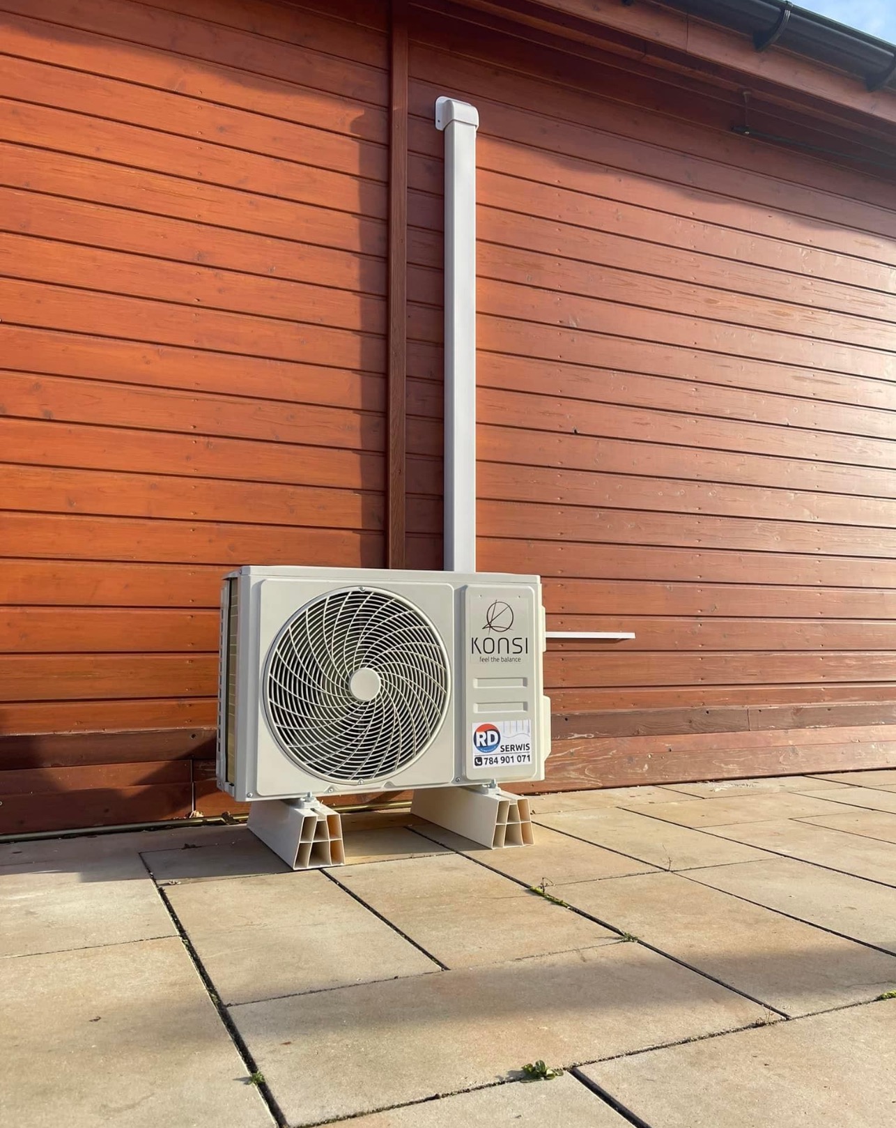 Zewnętrzna jednostka klimatyzacji marki Konsi zamontowana na elewacji budynku z drewnianą boazerią, widoczne białe rury odprowadzające skropliny oraz logo firmy serwisującej RD Serwis.