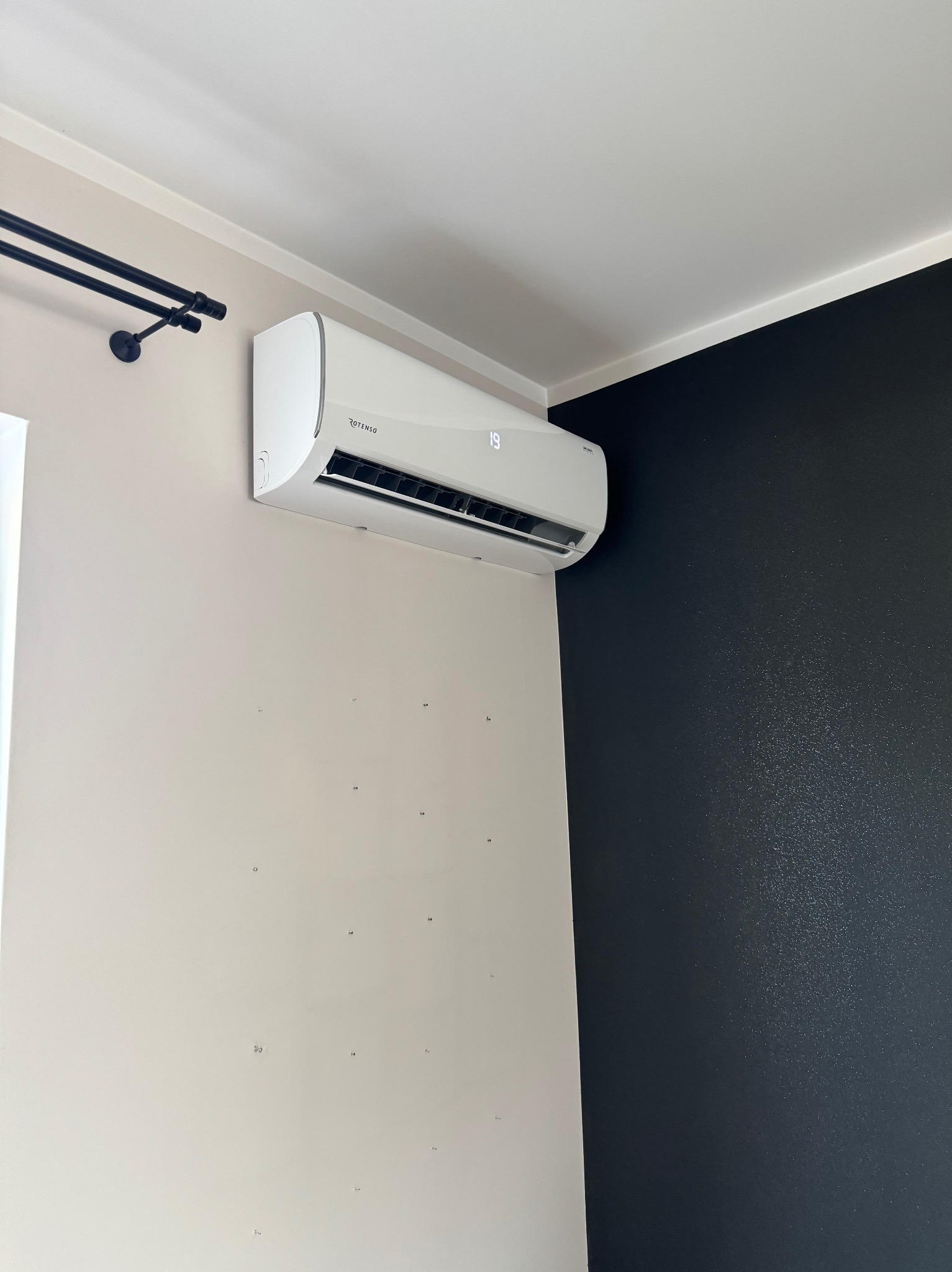 Biały klimatyzator Rotenso na ścianie w pokoju z beżową i czarną ścianą. Widoczny wyświetlacz temperatury. Minimalistyczny design i nowoczesne wykończenie wnętrza.