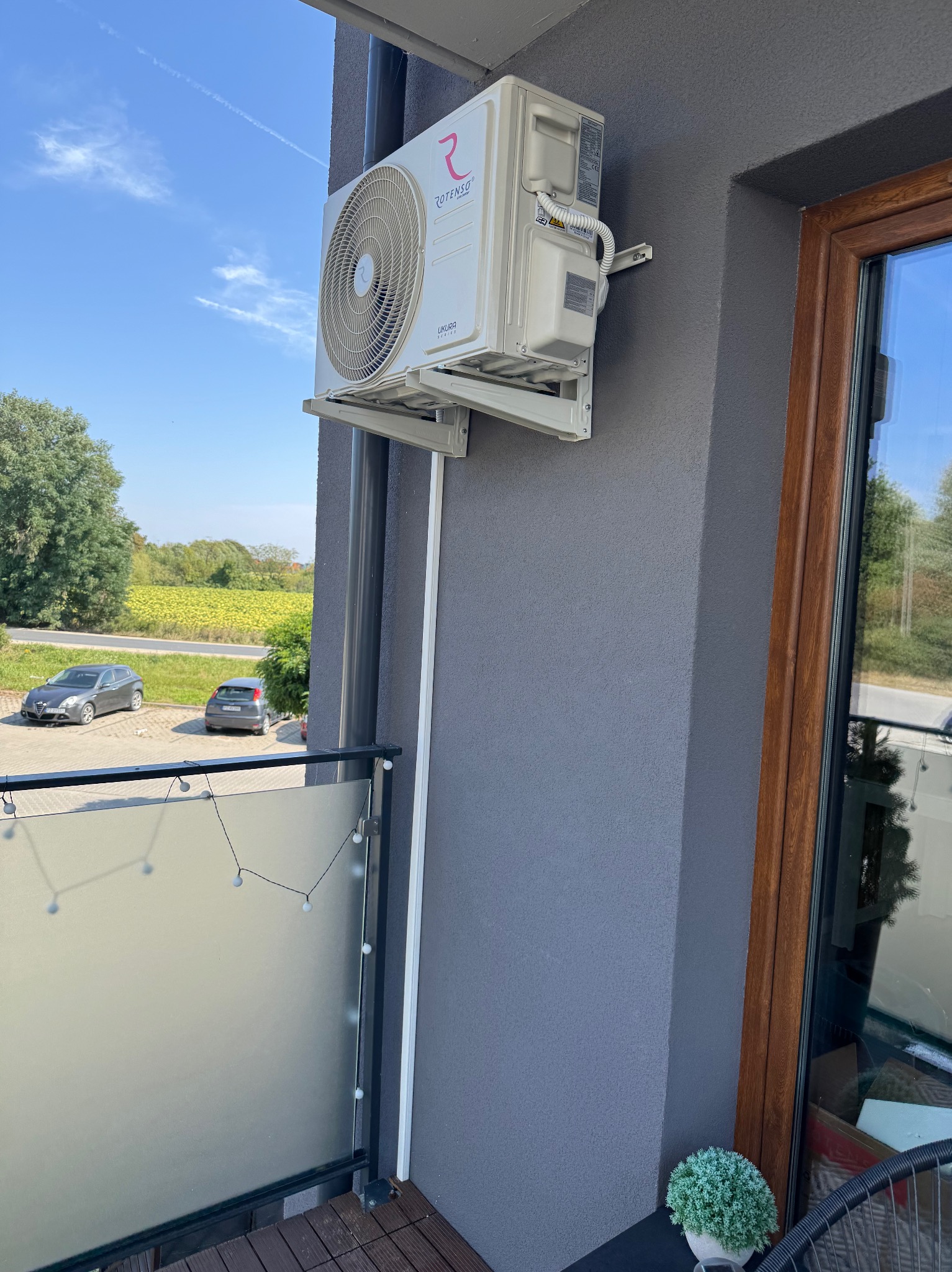 Biała klimatyzacja Rotenso Ukura Series zamontowana na szarej elewacji balkonu z widokiem na pole i niebo. Widoczne elementy instalacji.