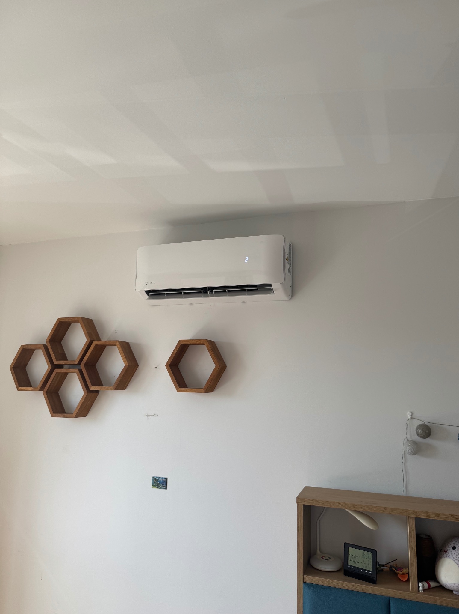 Biały klimatyzator na ścianie w pokoju, obok dekoracje w kształcie plastrów miodu. Widoczny fragment szafki nocnej z lampką i elektronicznym zegarem.