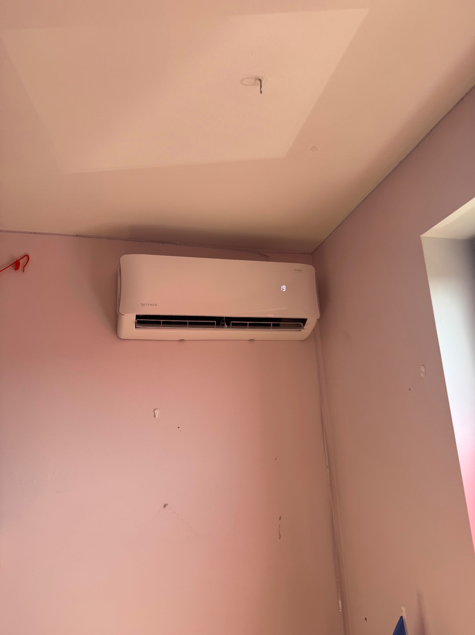 Biały klimatyzator Rotenso zamontowany na różowej ścianie w rogu pokoju, widoczny wyświetlacz z temperaturą 19 stopni Celsjusza. Ujęcie z dołu w kierunku sufitu.