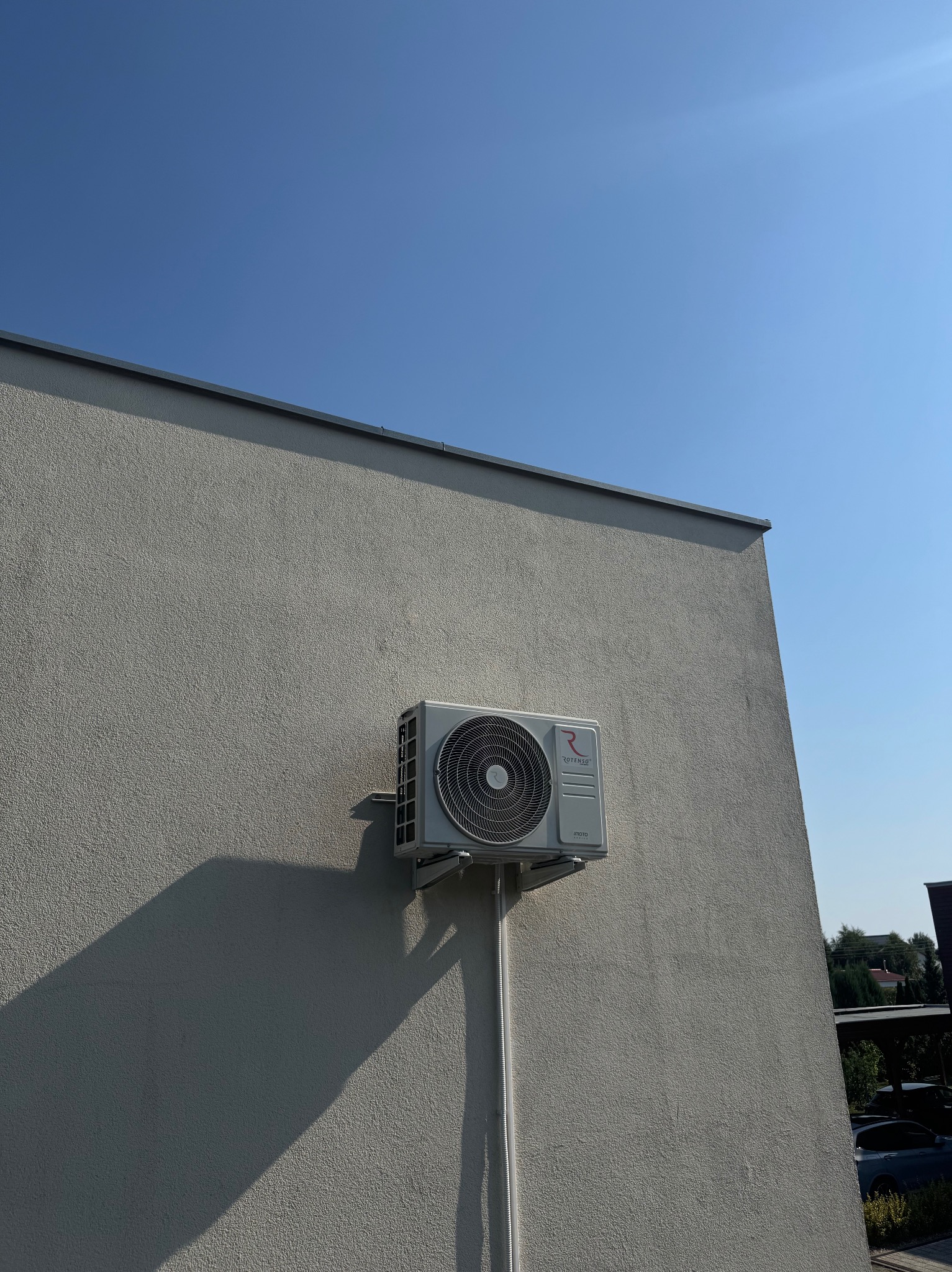 Biała jednostka zewnętrzna klimatyzacji marki Rotenso zamontowana na elewacji budynku pod błękitnym niebem. Widoczne rury instalacyjne i cień urządzenia.