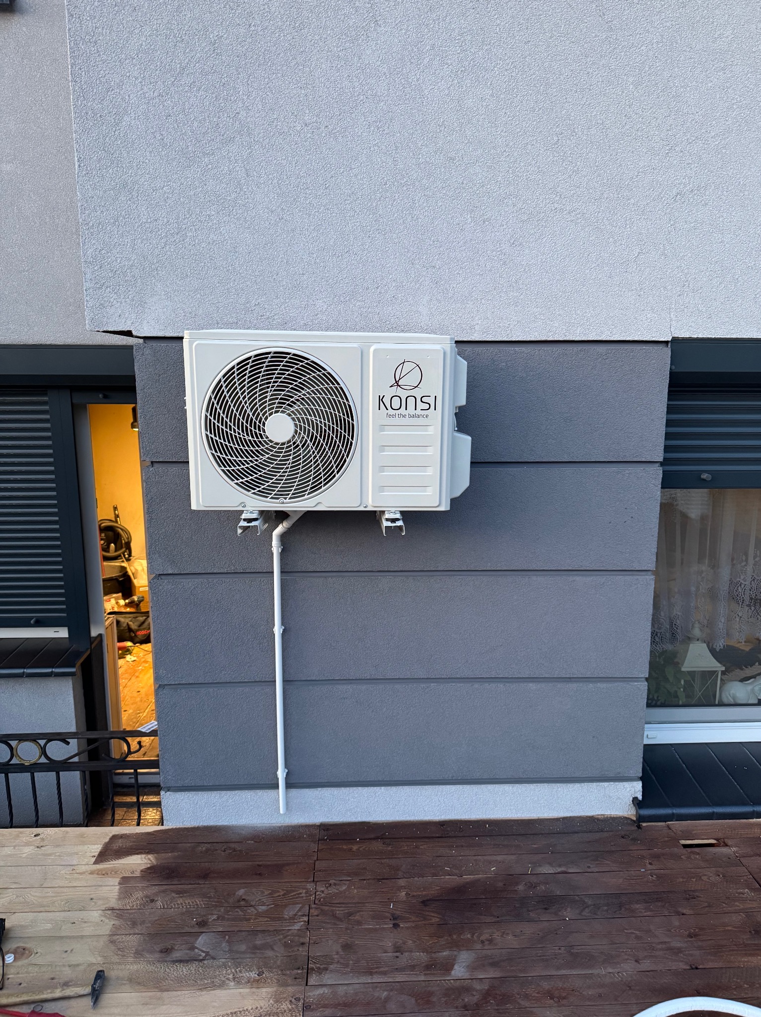 Biały klimatyzator marki Konsi zamontowany na szarej elewacji budynku, widoczny z zewnątrz. Na pierwszym planie drewniany taras w trakcie impregnacji.