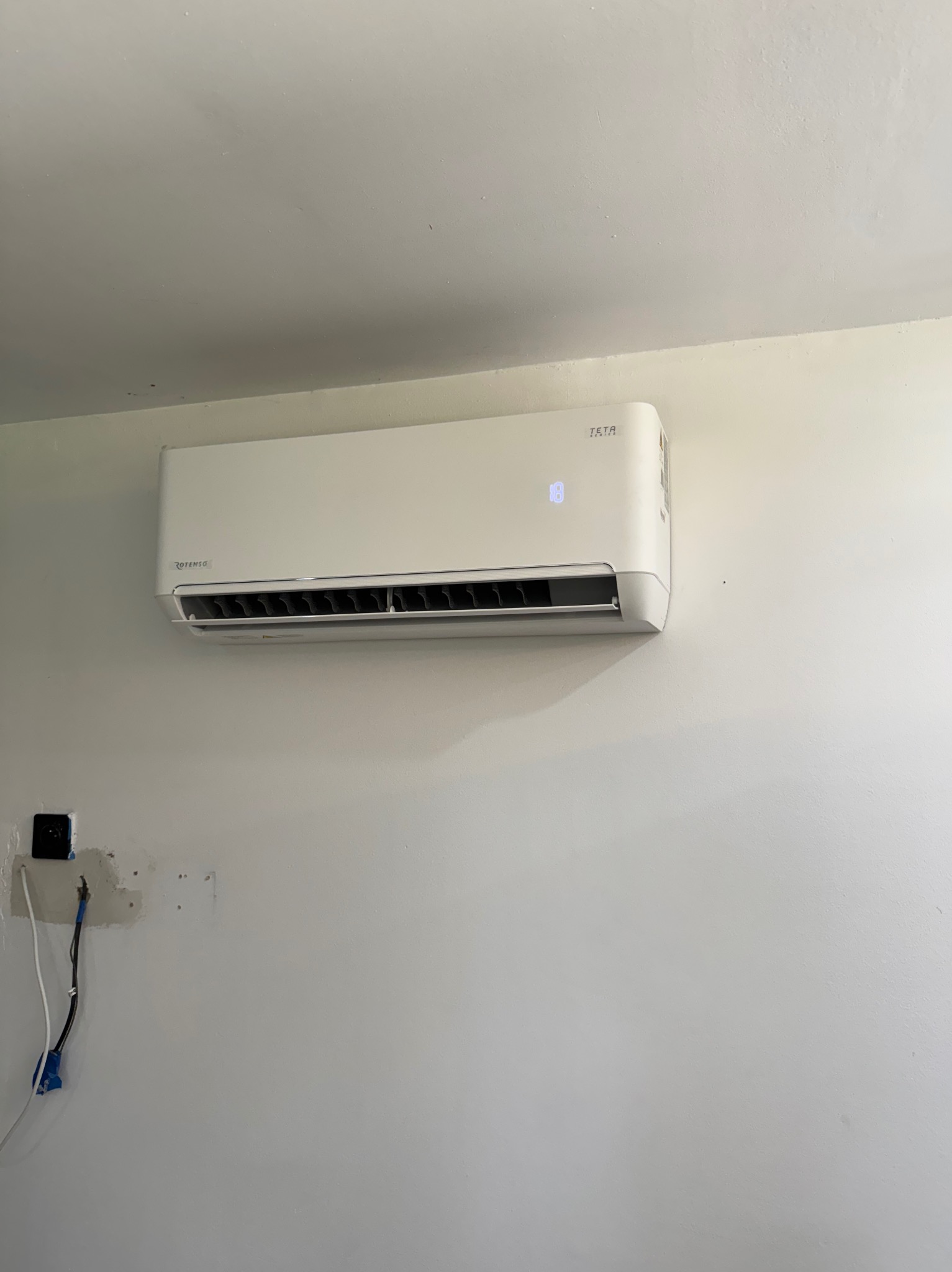Biała klimatyzacja TETA zamontowana na ścianie, widoczne kable wystające ze ściany, niedokończone wykończenie ściany przy gniazdku.