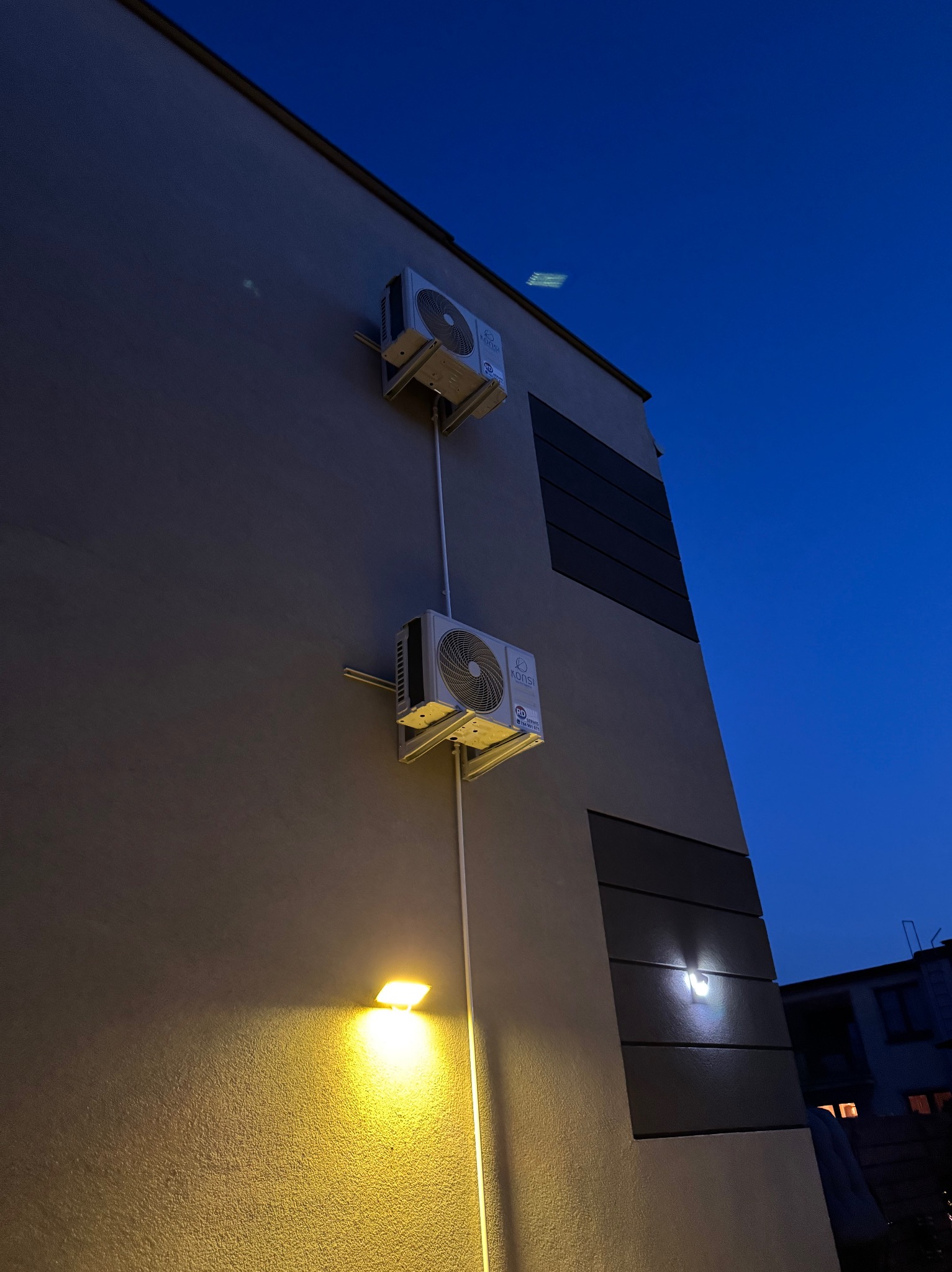 Dwa klimatyzatory zamontowane na elewacji budynku o zmierzchu, oświetlone lampą elewacyjną. Widoczne rury i przewody instalacyjne.