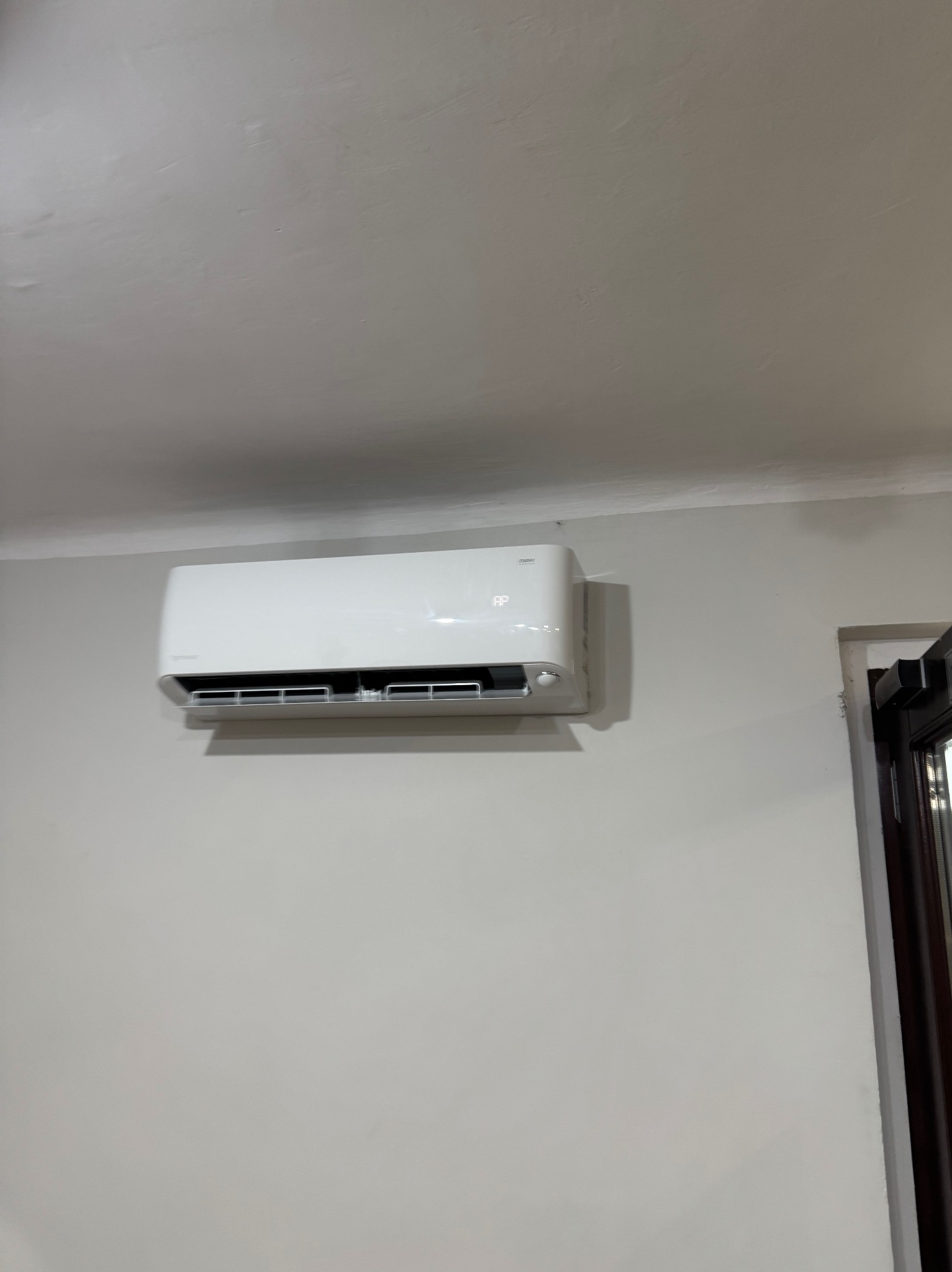 Biała klimatyzacja ścienna z widocznym wyświetlaczem, zamontowana na szarej ścianie w pomieszczeniu z fragmentem okna po prawej stronie.