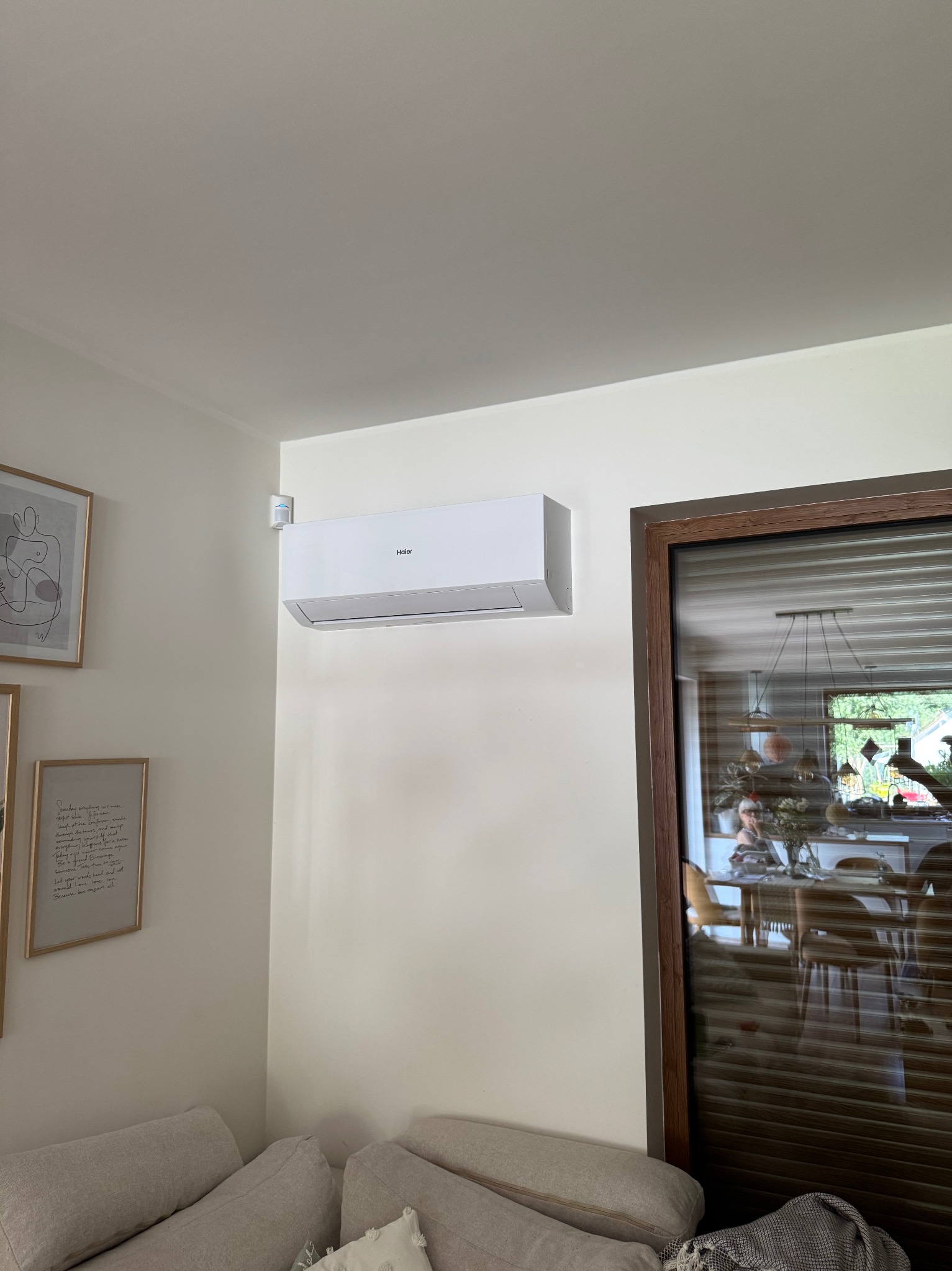 Biała klimatyzacja Haier na ścianie w jasnym wnętrzu, widoczny fragment sofy i dekoracje ścienne. Ujęcie z dołu, naturalne oświetlenie.