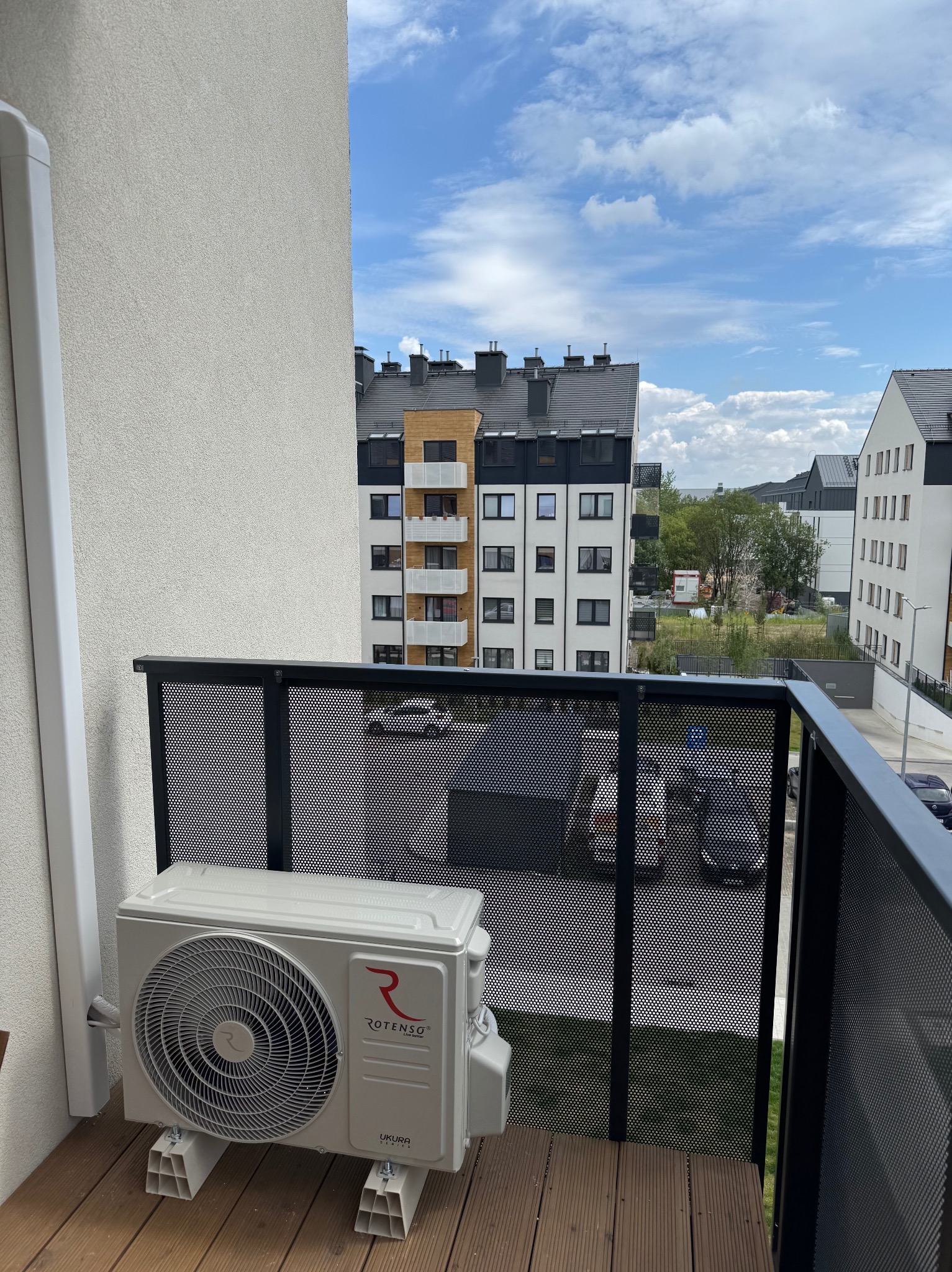 Biały klimatyzator Rotenso Ukura na balkonie z drewnianą podłogą i czarną balustradą na tle osiedla mieszkaniowego w słoneczny dzień.
