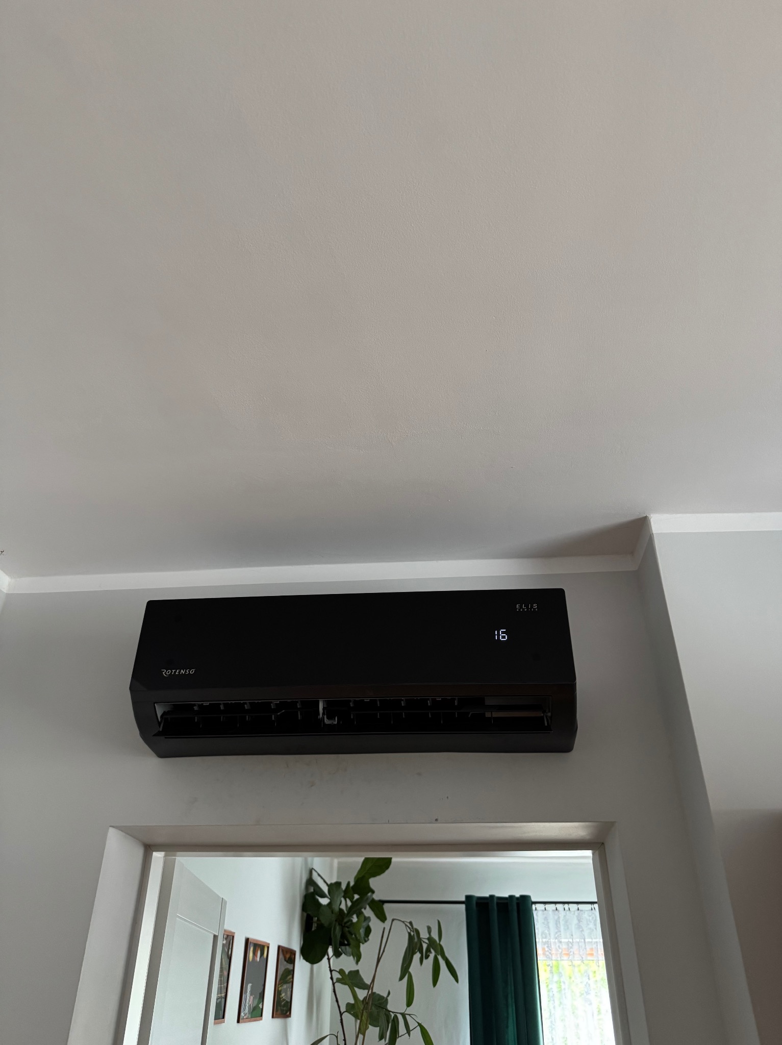 Czarna klimatyzacja Rotenso Elis zamontowana na ścianie nad drzwiami, widoczny wyświetlacz z temperaturą 16 stopni, wnętrze pokoju z roślinami w tle.