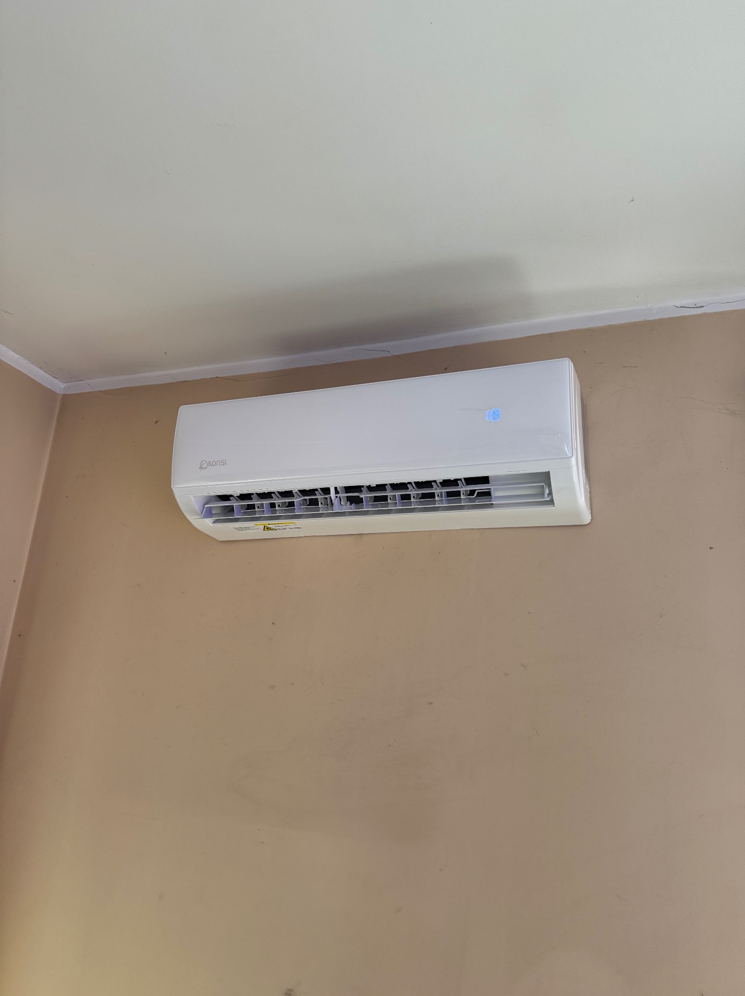 Biała klimatyzacja marki Konsi zamontowana na beżowej ścianie, widoczny wyświetlacz z temperaturą 18 stopni, kątowy widok z dołu.