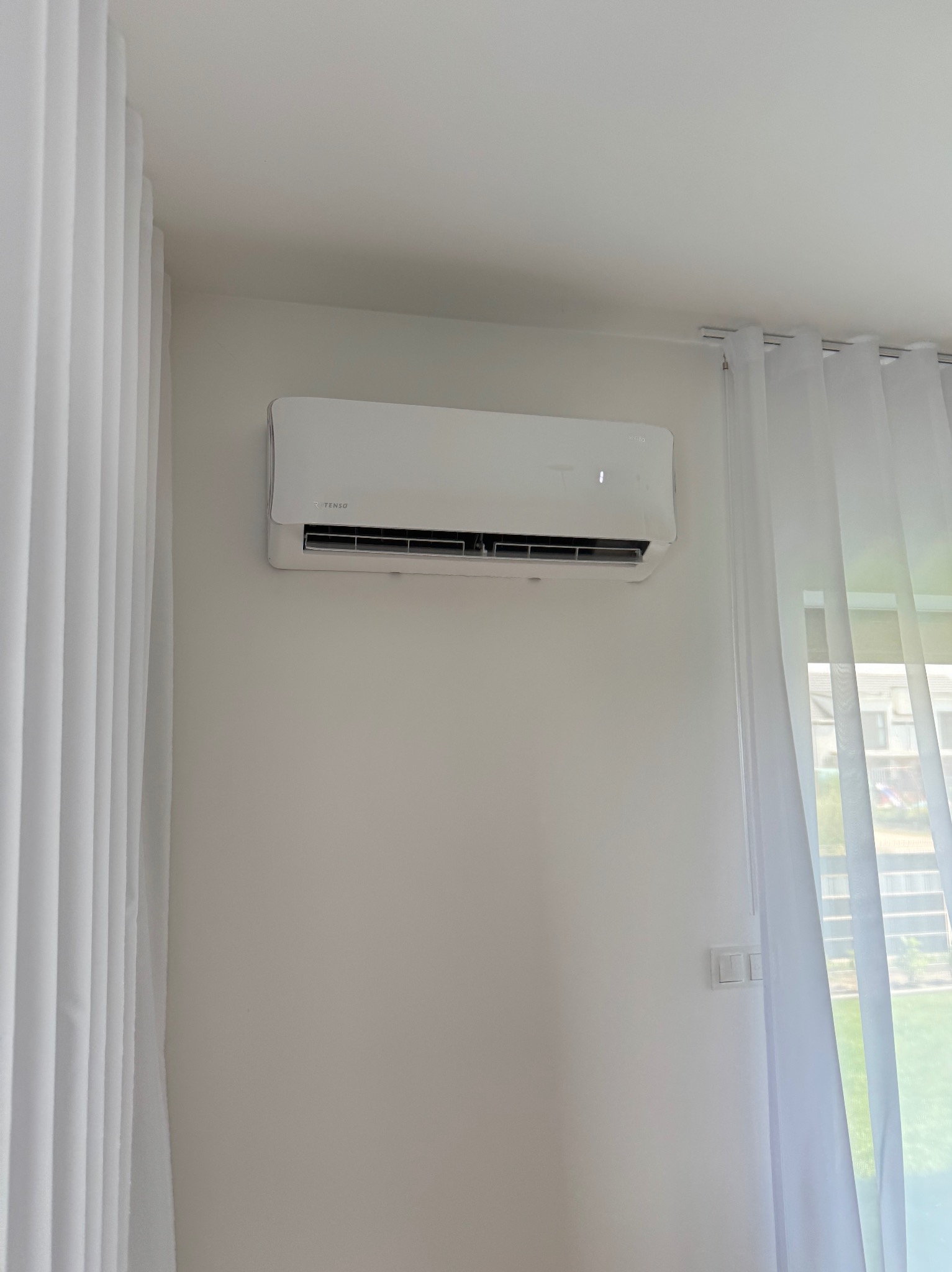 Biała klimatyzacja ścienna Tenso w minimalistycznym wnętrzu z białymi zasłonami. Widoczny fragment okna i zieleni na zewnątrz. Czysty montaż.
