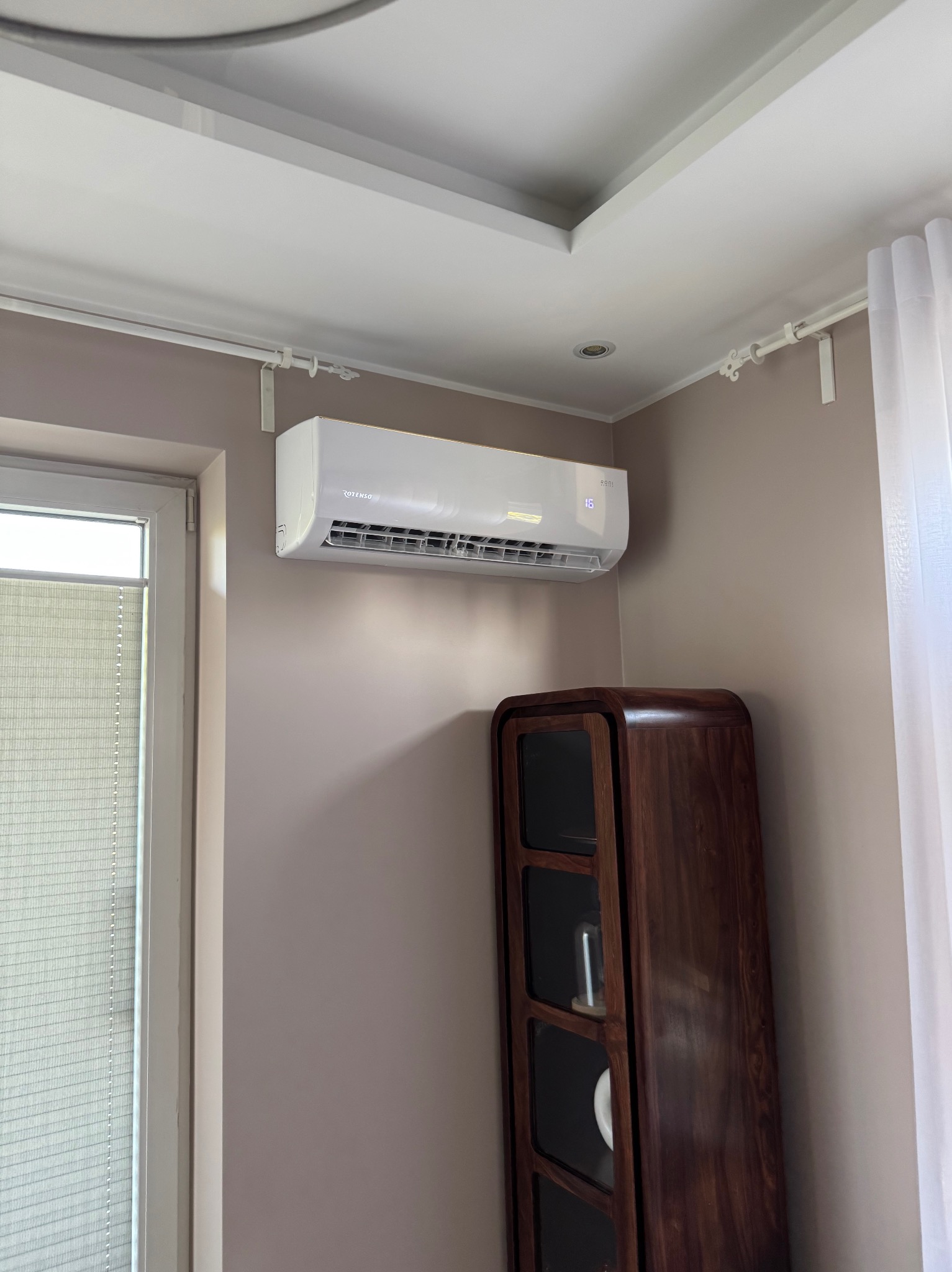 Biały klimatyzator Rotenso na ścianie w pokoju, obok okna i drewnianej witryny. Widoczny wyświetlacz z temperaturą 16 stopni.