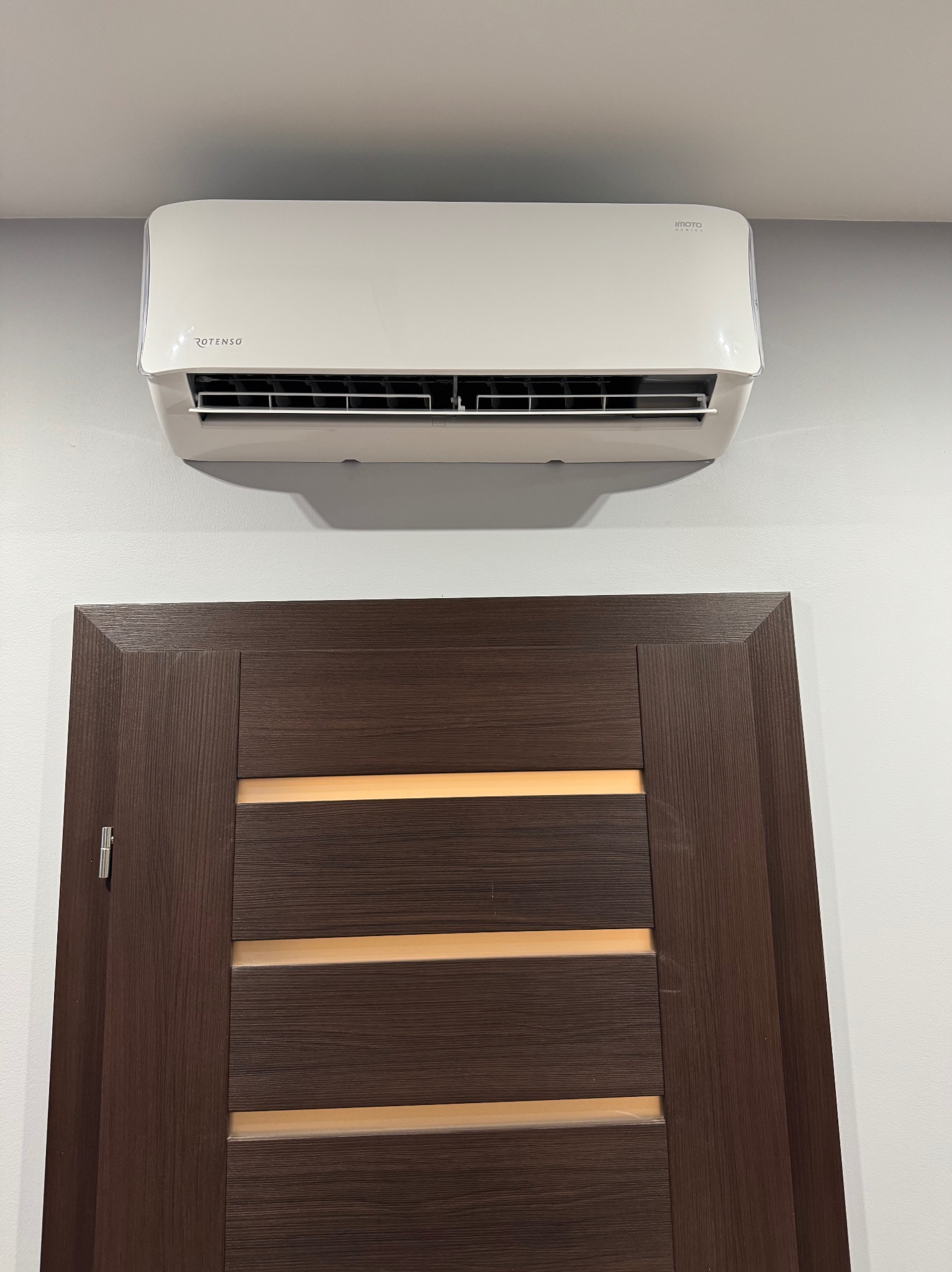 Biała klimatyzacja ROTENSO zamontowana nad brązowymi drzwiami z poziomymi przeszkleniami na jasnej ścianie. Widok z dołu.