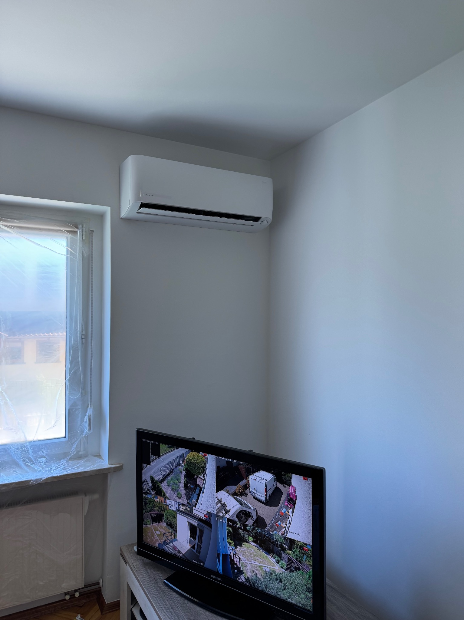 Biała klimatyzacja ścienna nad telewizorem z widokiem z kamery. Okno zabezpieczone folią malarską. Jasne wnętrze, minimalistyczny design.