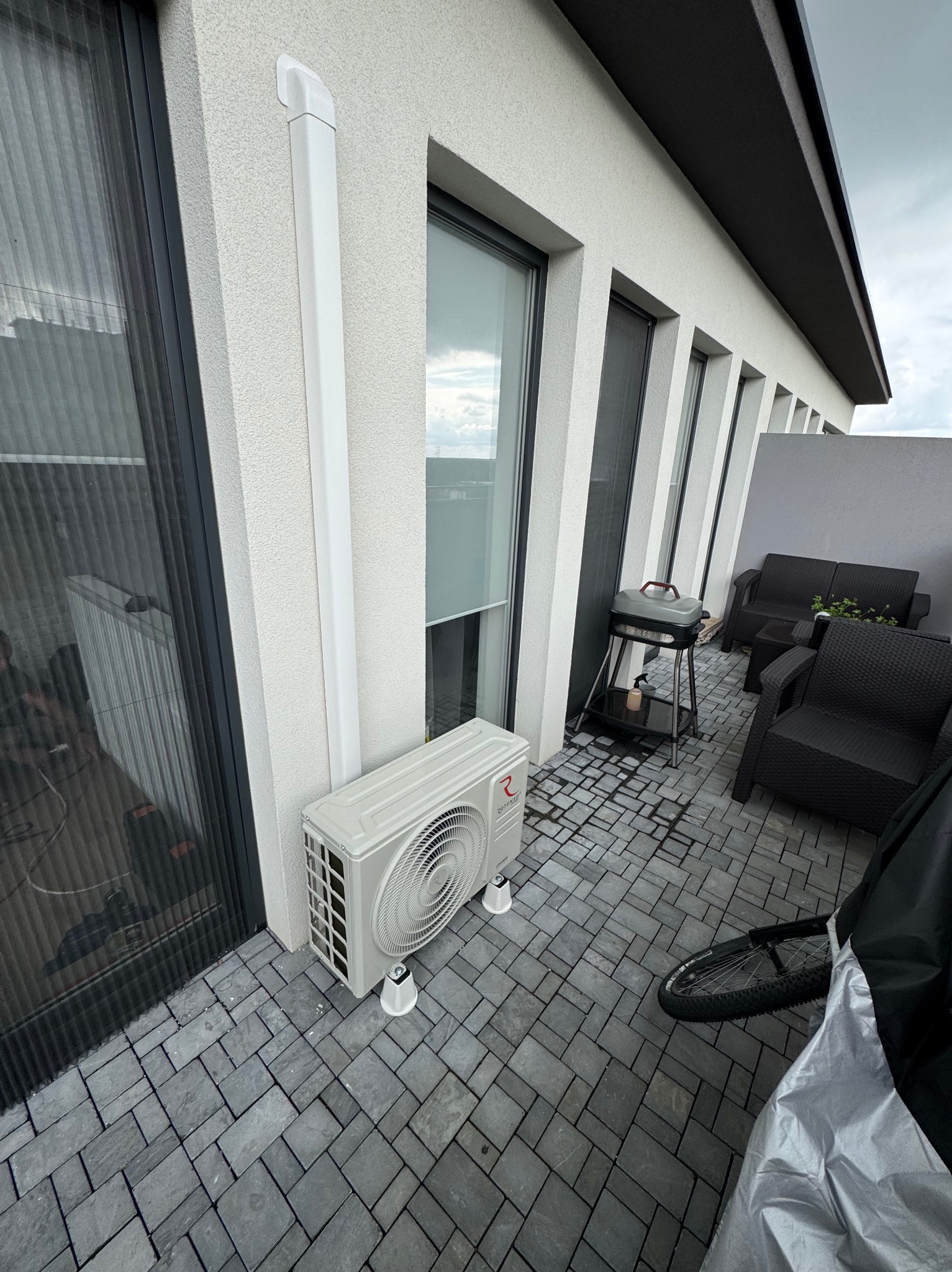Biała jednostka zewnętrzna klimatyzacji na balkonie z szarą kostką brukową, widoczna rura odprowadzająca skropliny, meble ogrodowe w tle.