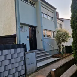 Odnowiona elewacja dwupiętrowego domu w Sławie, z numerem 21 nad ciemnymi drzwiami wejściowymi, szare schody z metalową barierką, widoczne okna z roletami i fragment ogrodzenia.