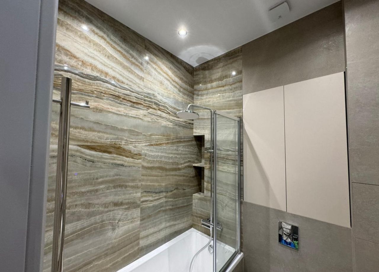 Wykończona łazienka z wanną i kabiną prysznicową, ściany wyłożone płytkami imitującymi kamień z wyraźnymi warstwowymi wzorami. Minimalistyczne szafki w tle.