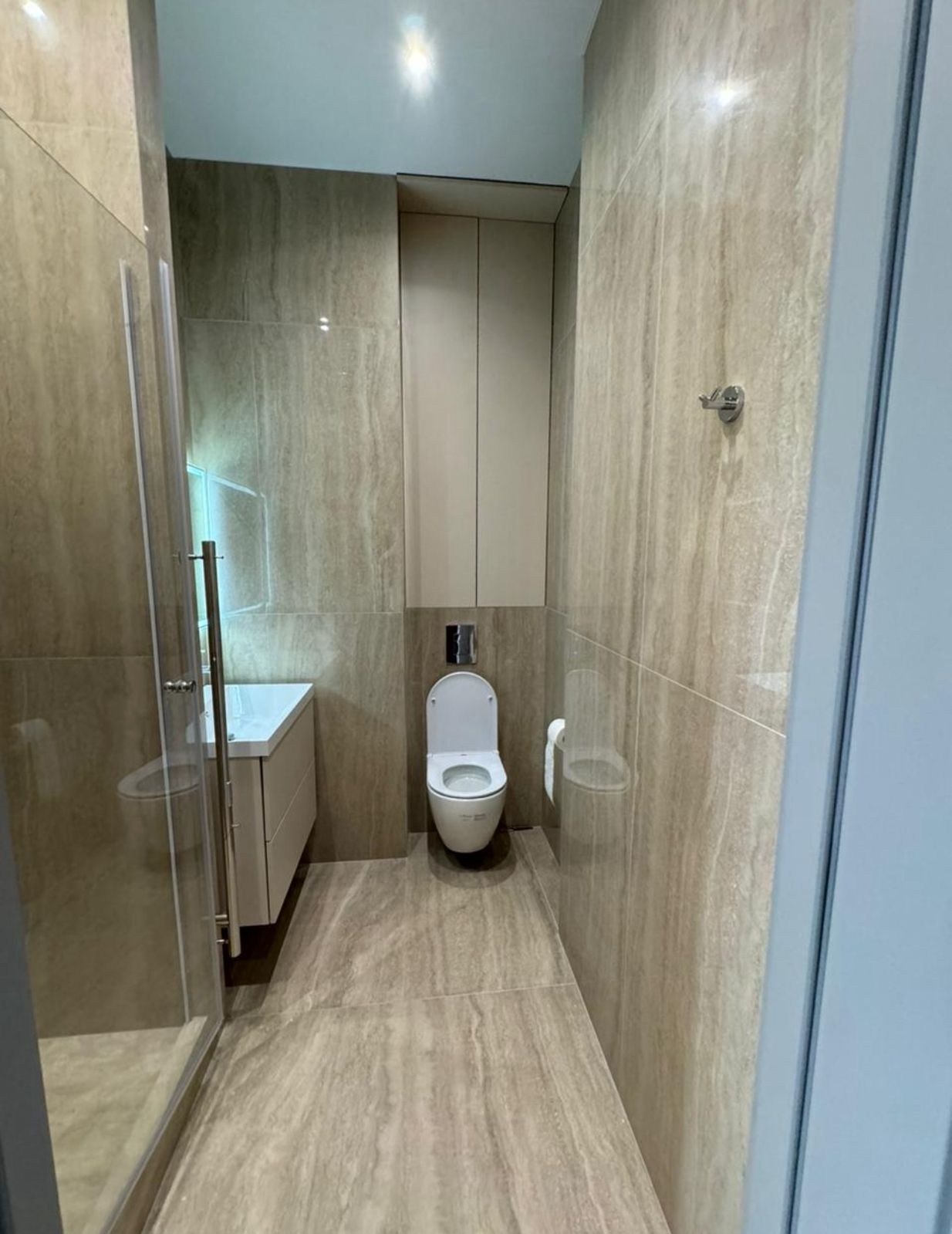 Wykończona łazienka z kabiną prysznicową, toaletą i szafką pod umywalką. Płytki imitujące marmur na ścianach i podłodze. Minimalistyczny design, neutralna kolorystyka.