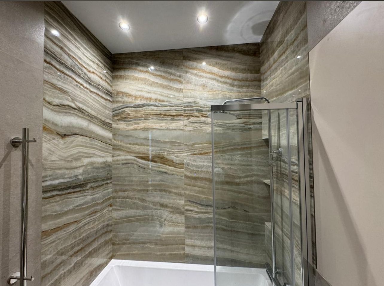 Wykończona łazienka z kabiną prysznicową i płytkami imitującymi onyks na ścianach. Widoczne oświetlenie sufitowe i chromowany wieszak na ręczniki.